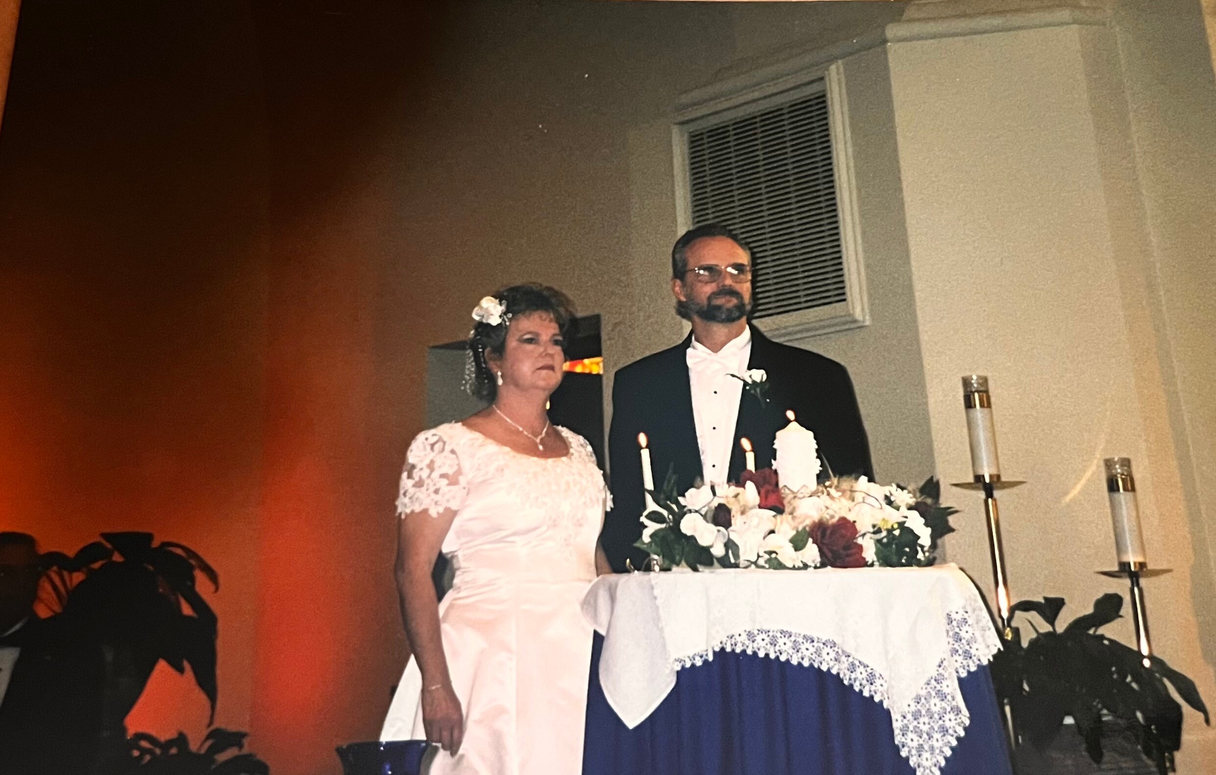 Bride in a white lace wedding dress stands beside a groom in a tuxedo at a wedding cake with candles and flowers on the table.