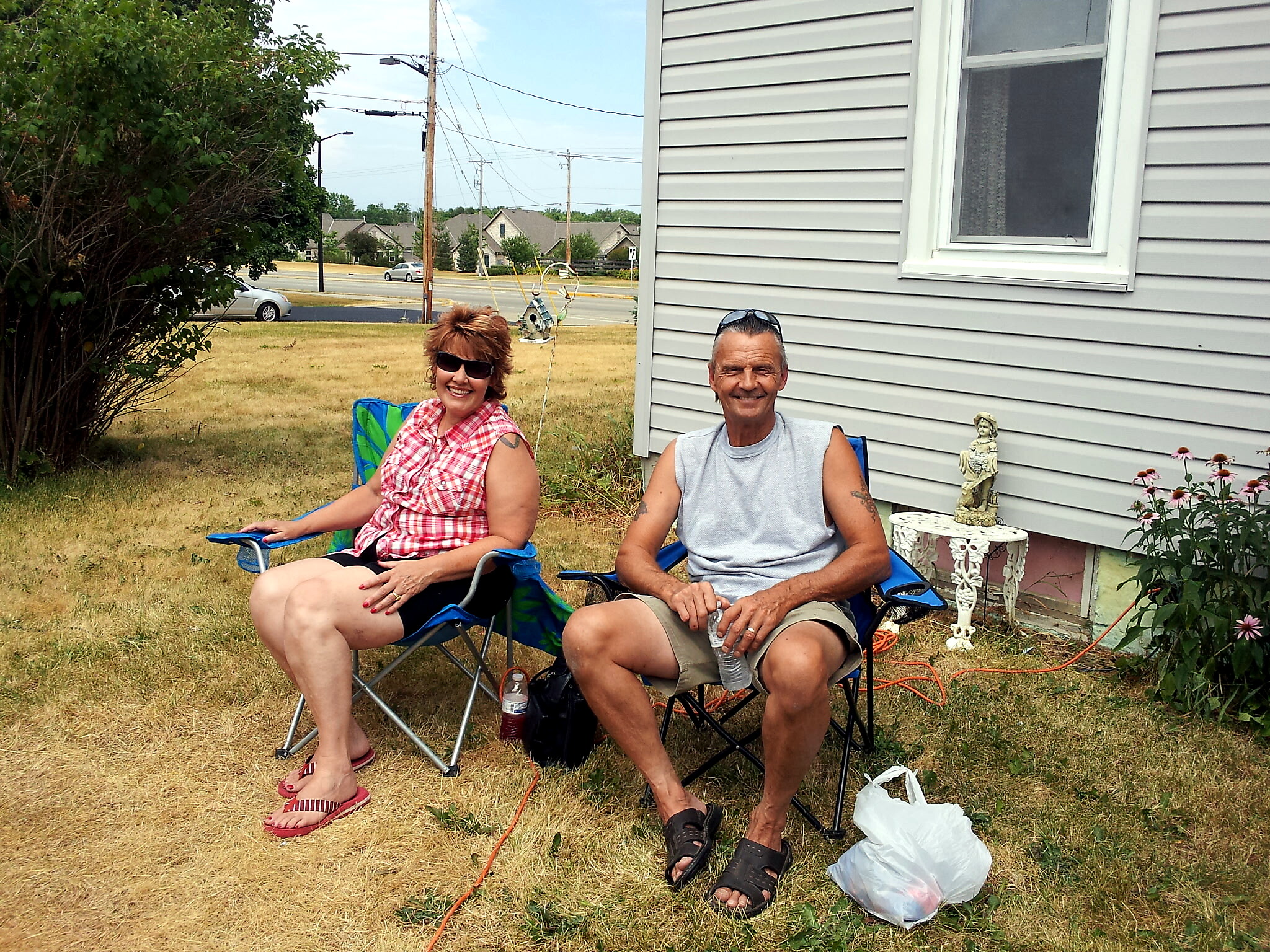 Two smiling adults sit in blue lawn chairs on a dry, brown yard beside a gray house, with a street and power lines in the background.