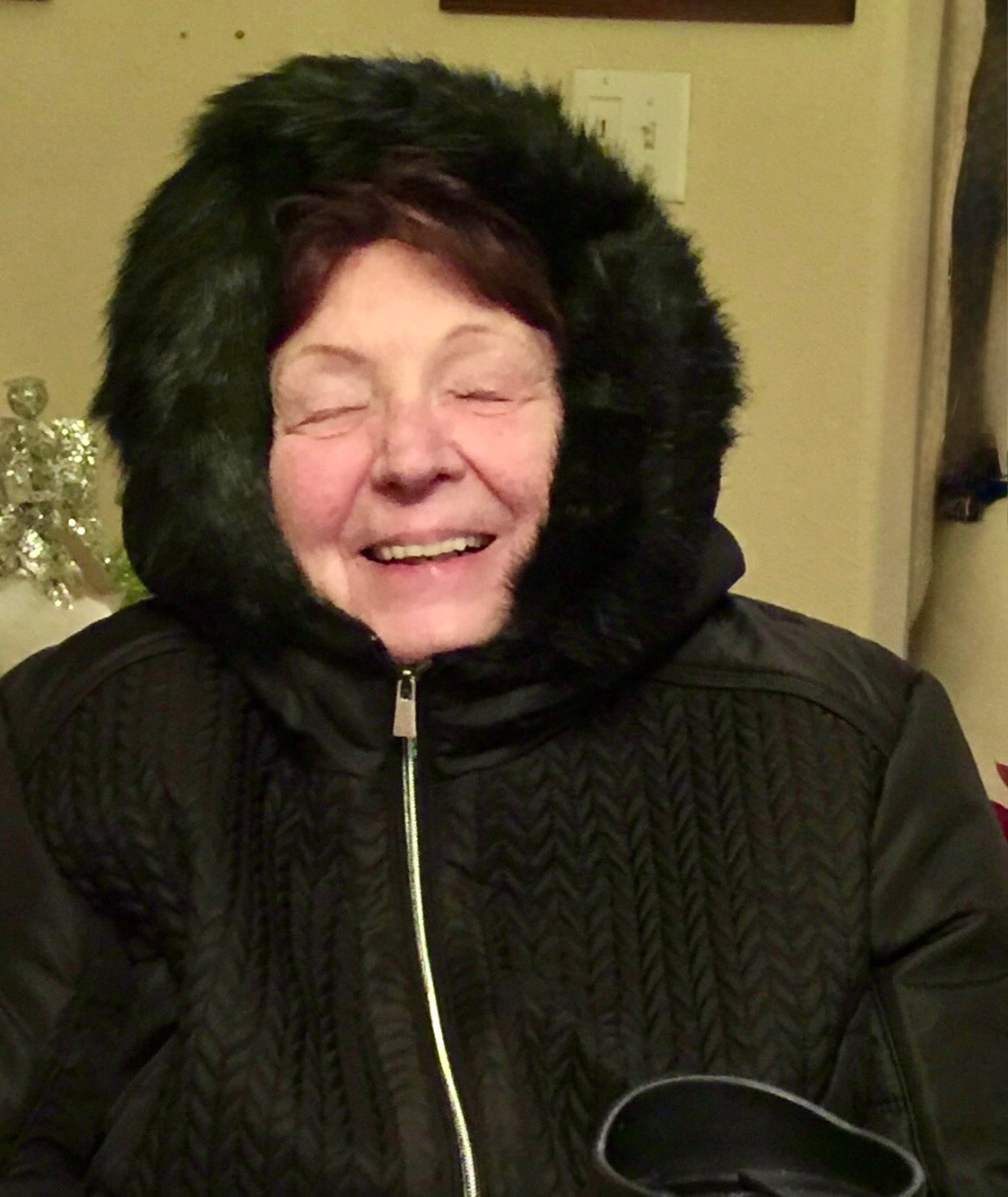 Older woman smiling, wearing a black hood with faux fur trim and a zipped black coat indoors.