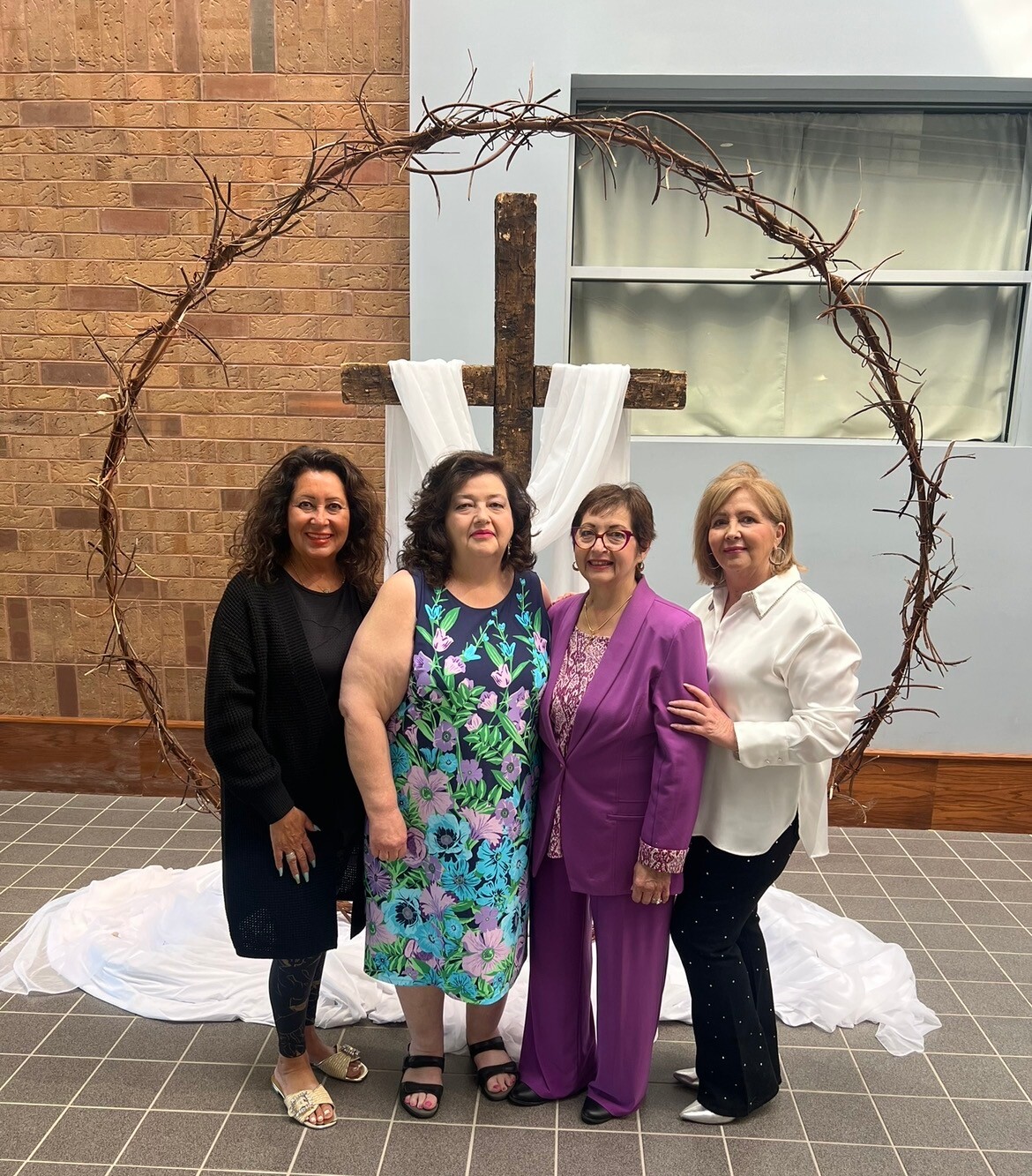 Four women stand together posing in front of a rustic wooden cross with a white fabric drapery arch outdoors/indoors, smiling at the camera.
