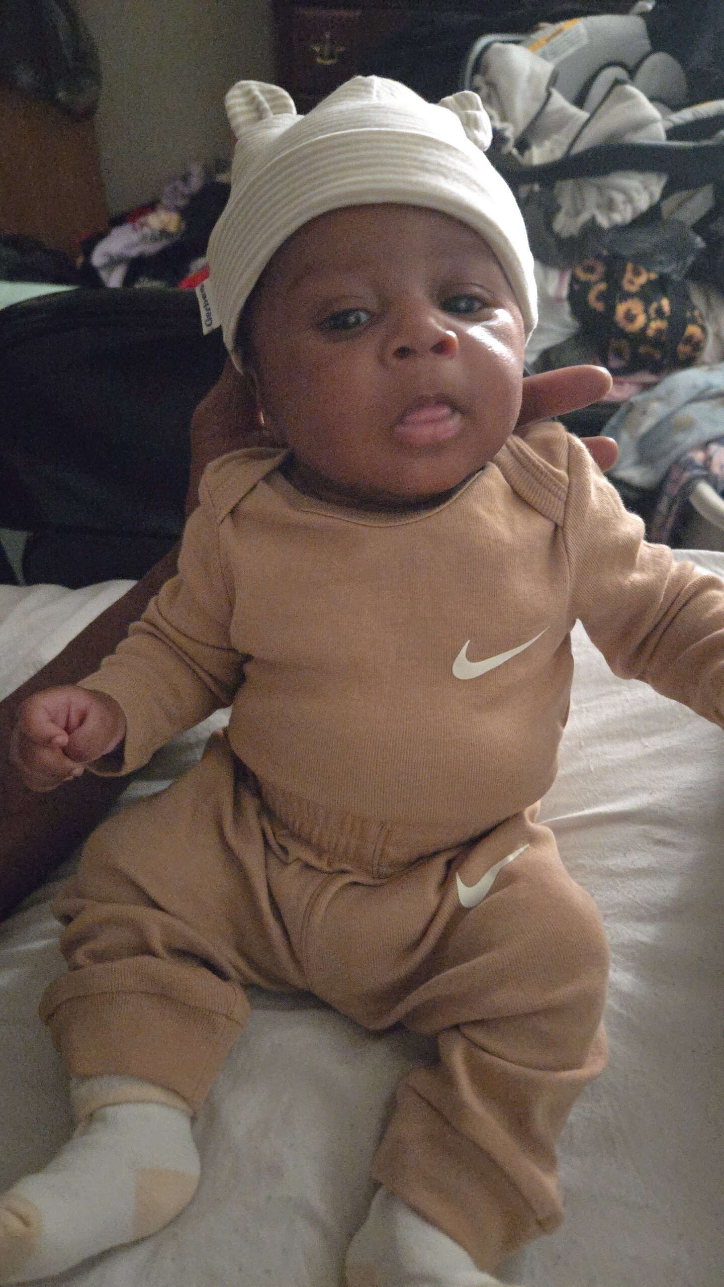 Baby in a tan Nike outfit with a white bear-ear hat, lying on a bed and looking at the camera.