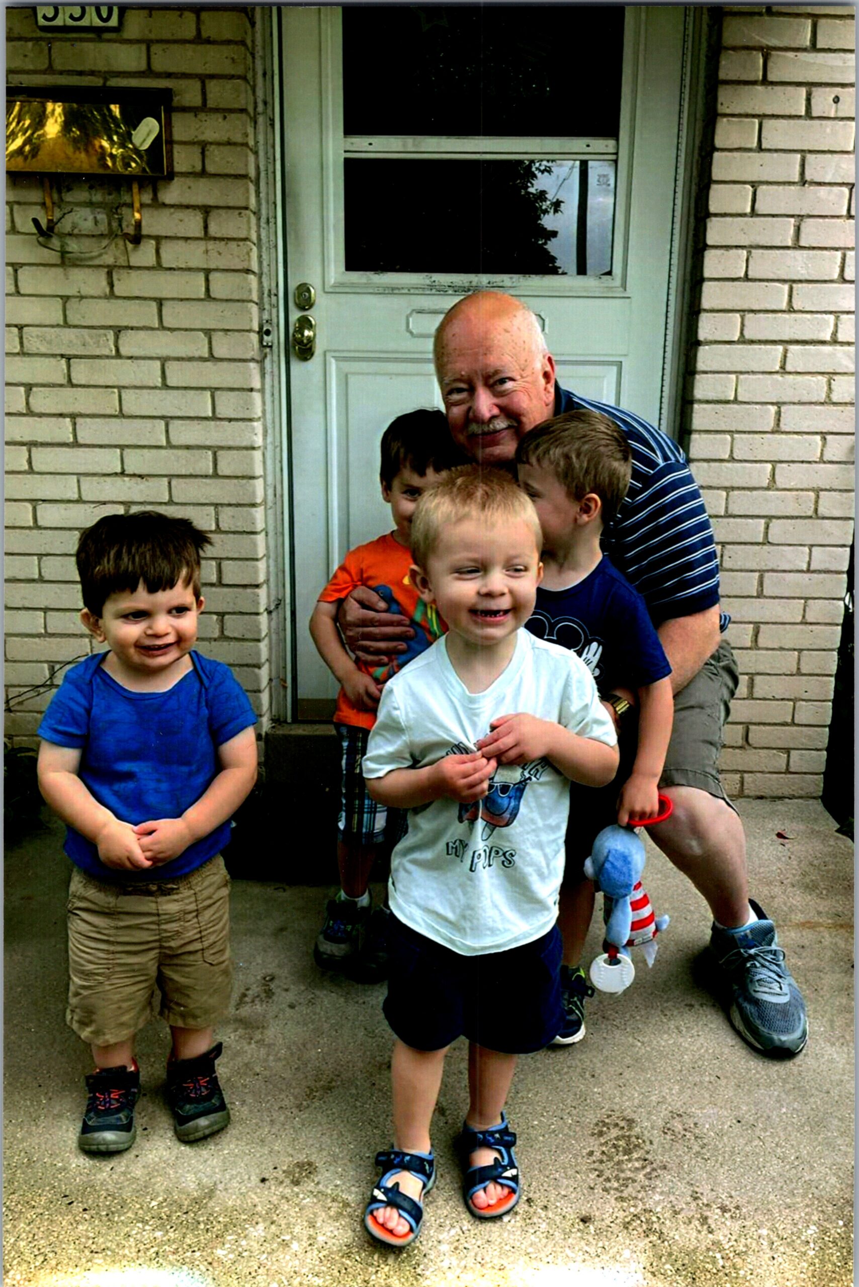 Older man in a striped shirt with several smiling children posing on a brick porch in front of a pale door.