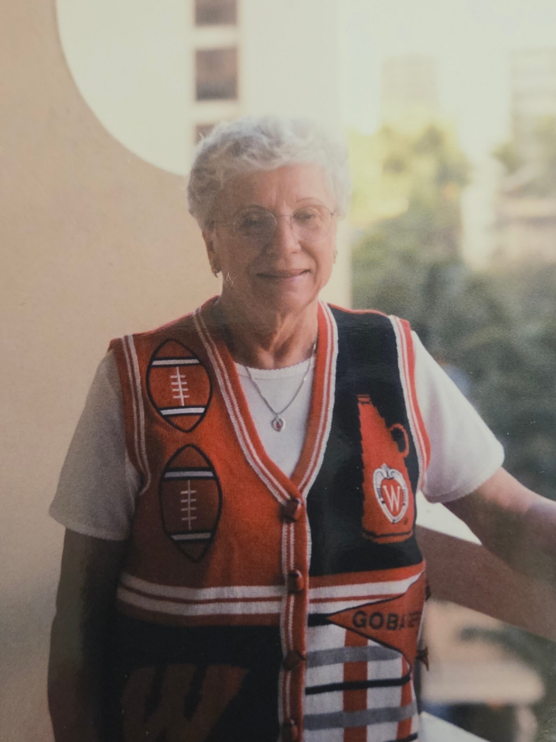 Older woman with short gray hair and glasses smiling, wearing a red and orange knit vest with football patches.