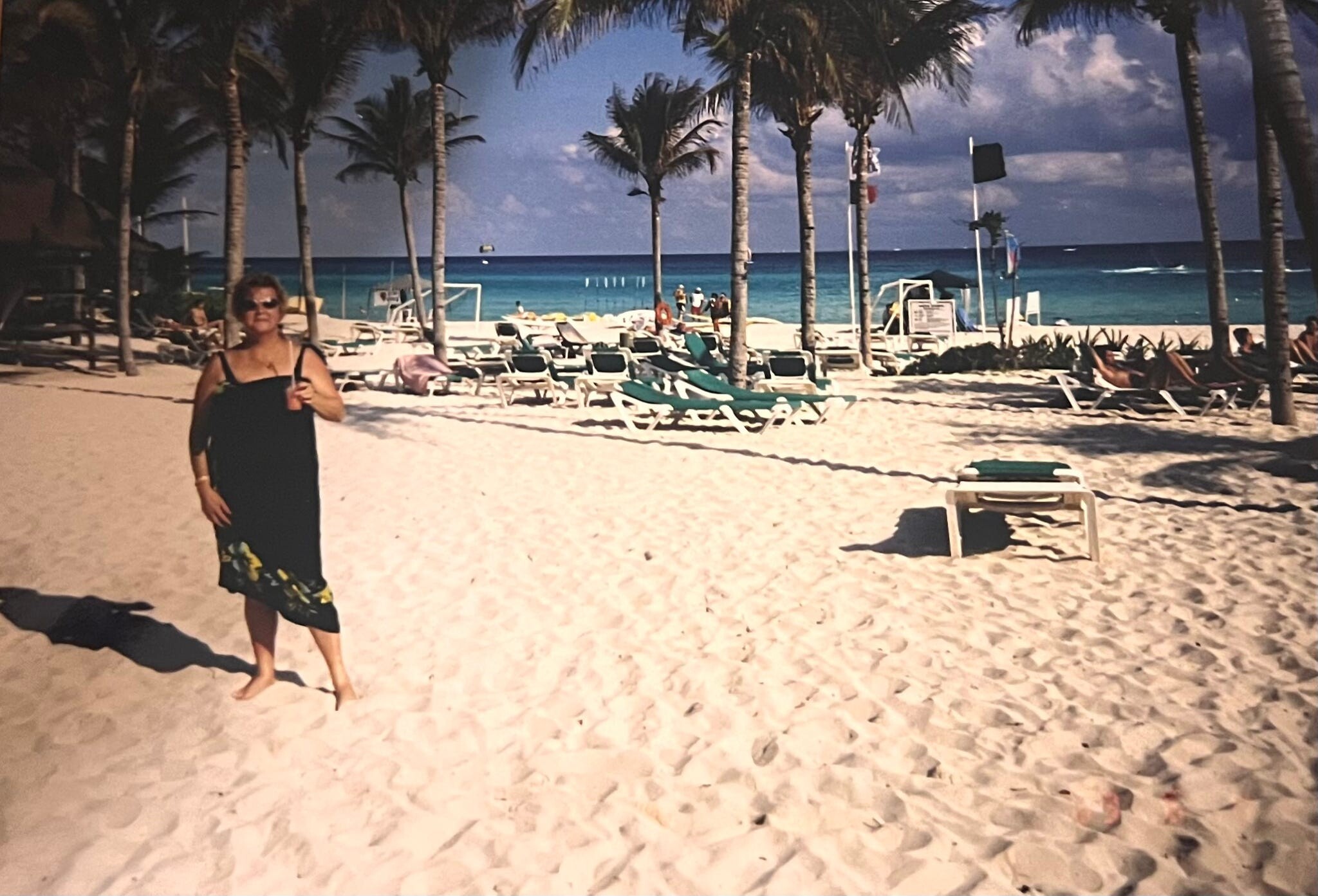 Woman in a black dress stands on a sunny sandy beach with palm trees and blue-green ocean in the background.