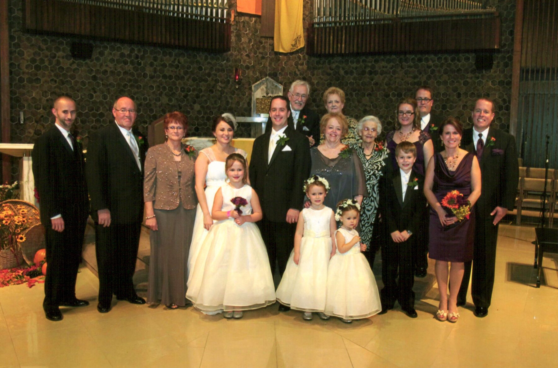 Wedding party posing in a church: bride and groom center, bridesmaids and flower girls in white dresses, family and guests in formal attire gathered around them at the altar area.