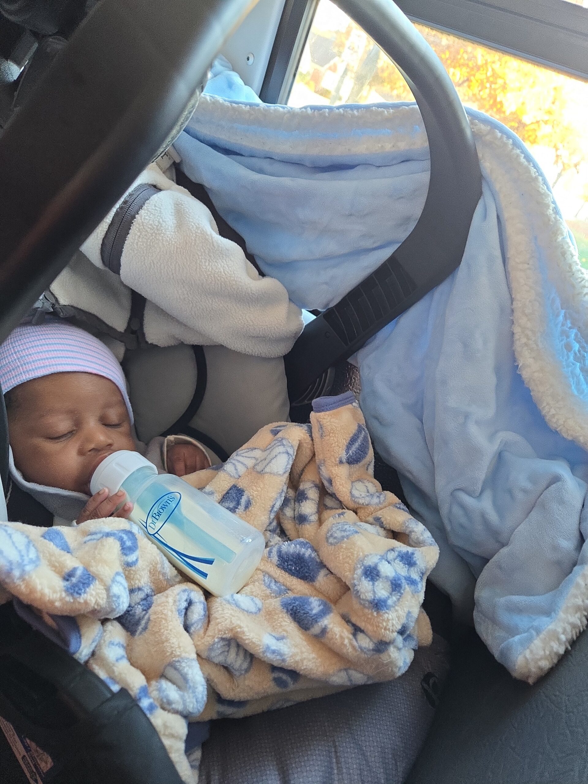 Sleeping infant in a car seat, wrapped in blue fleece and a soft blanket, drinking from a bottle.