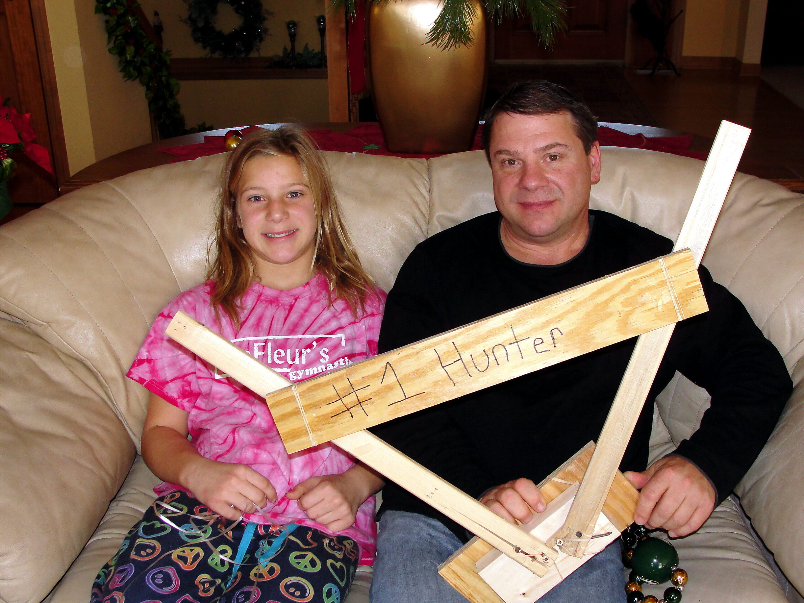Two people sit on a beige couch with a wooden DIY cross held in front of them, smiling at the camera.