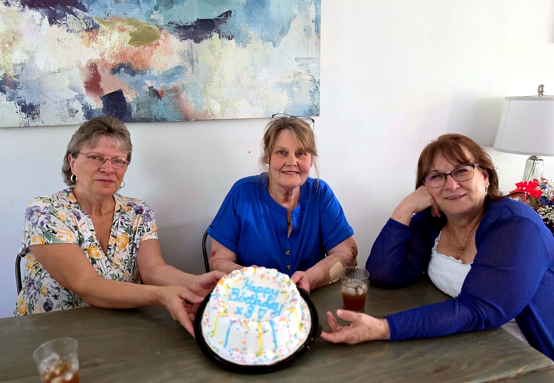 Three women sit at a table indoors, celebrating a birthday with a colorful frosted cake in the foreground.