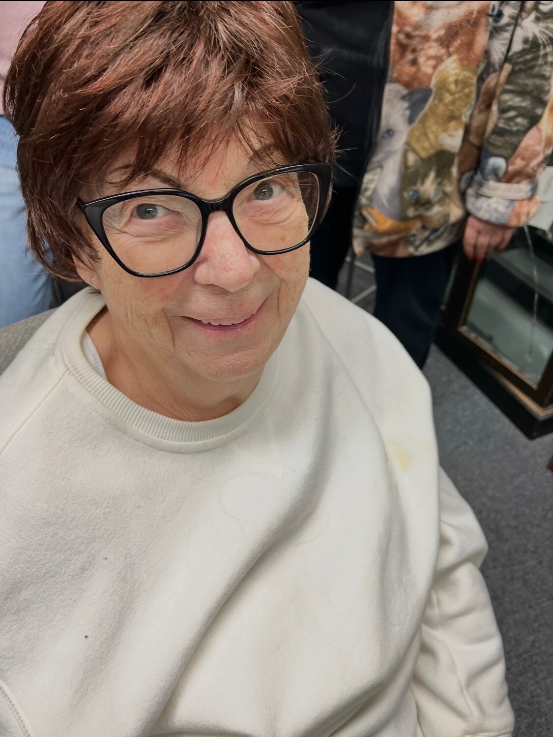 Close-up of an elderly woman with short brown hair and black-framed glasses smiling at the camera.