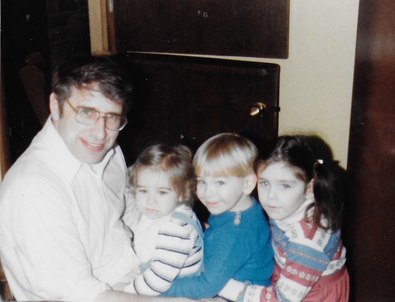 Father and three children posing close together beside a dark fireplace, smiling at the camera. The man wears glasses and a light shirt; the kids wear striped, blue, and patterned sweaters.