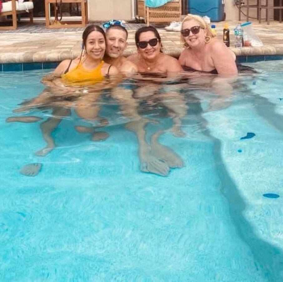 Four people posing in a pool, smiling at the camera with a poolside patio in the background.