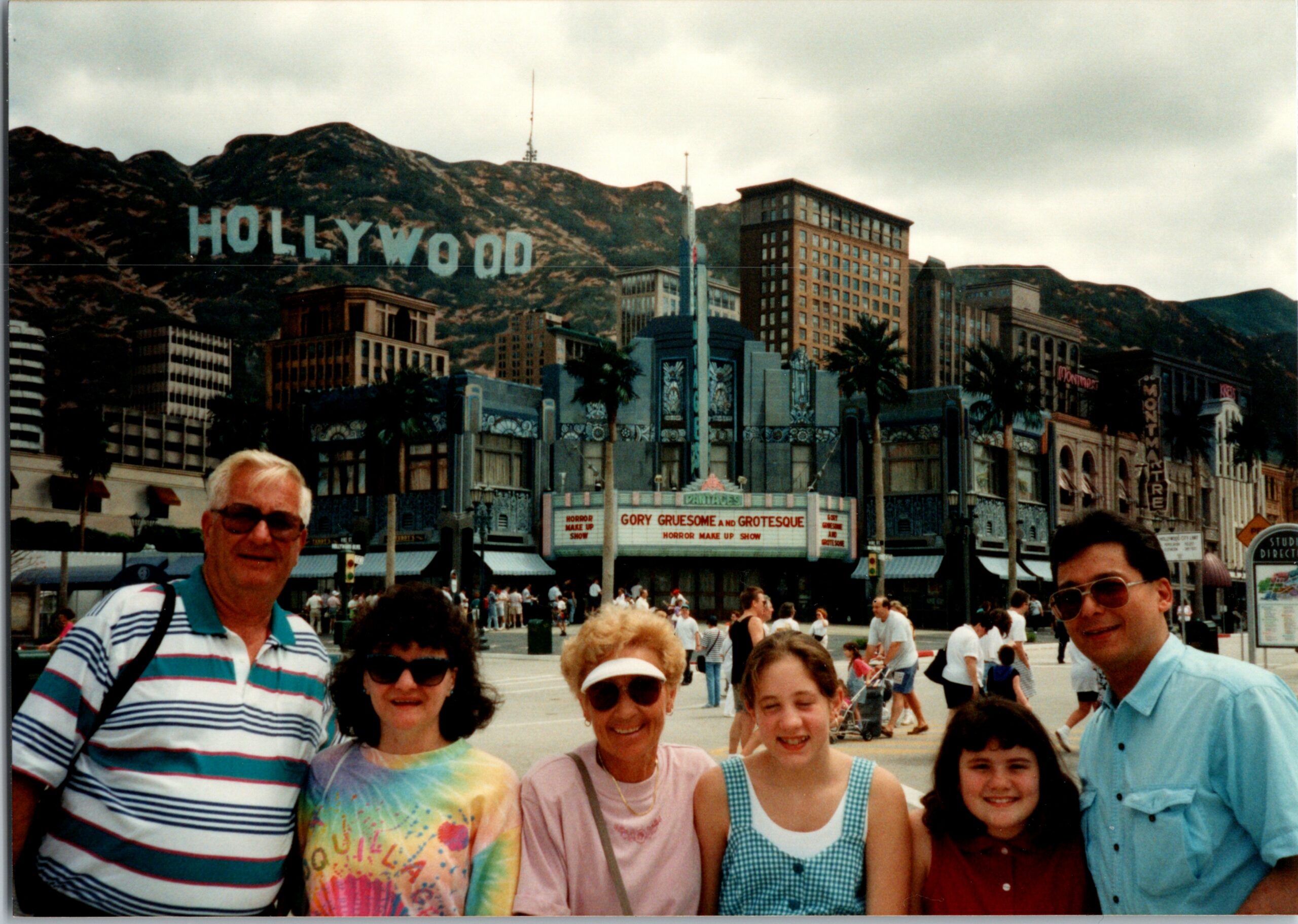 Group of smiling tourists posing for a photo on a sunny street with the Hollywood sign on the hills in the background.
