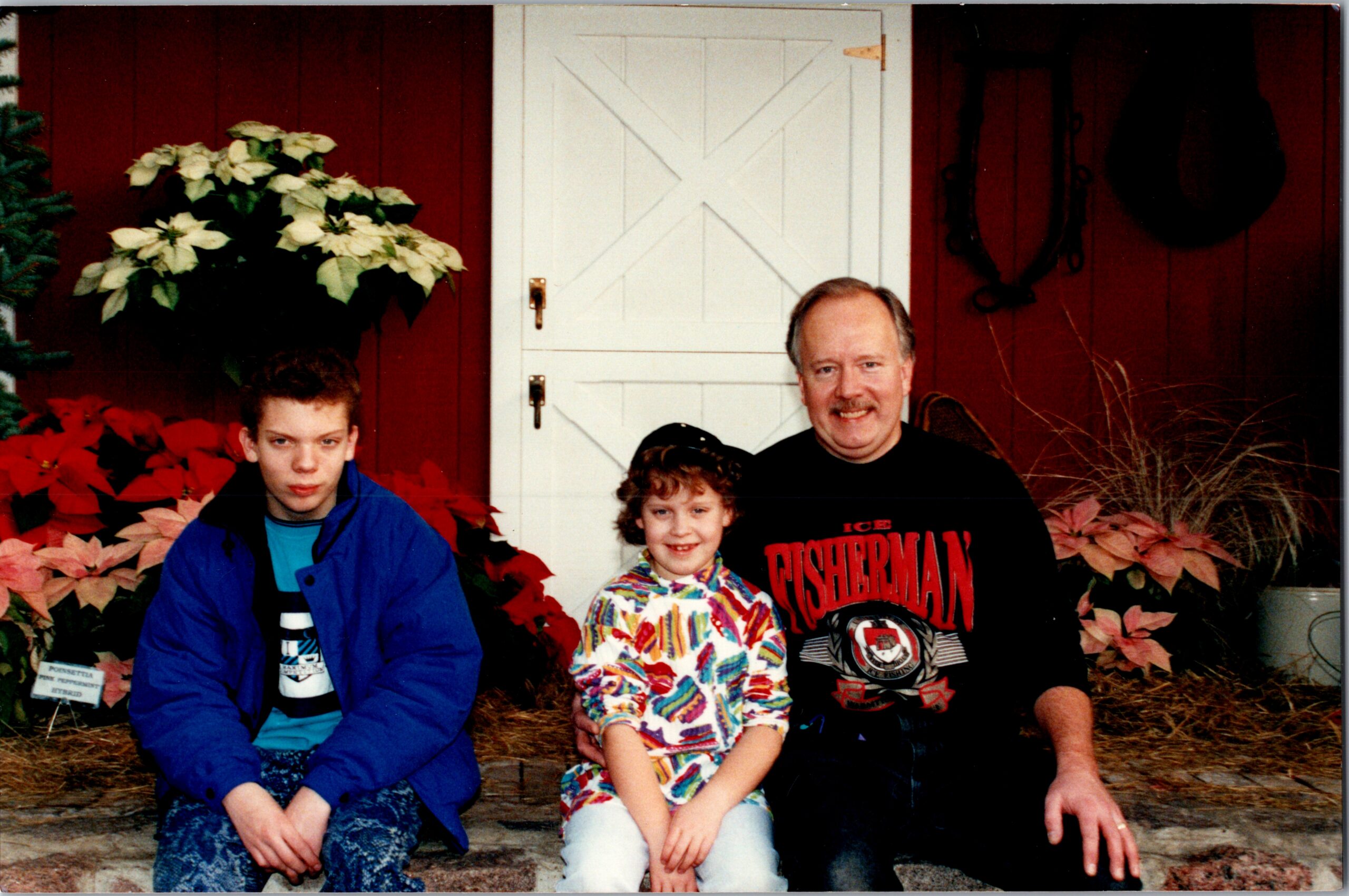 A family of four sits on the front steps in front of a white paneled door, with red siding and holiday plants (poinsettias) on both sides; a man wearing a black shirt sits next to a woman or child on colorful clothing, while two younger kids pair with them.