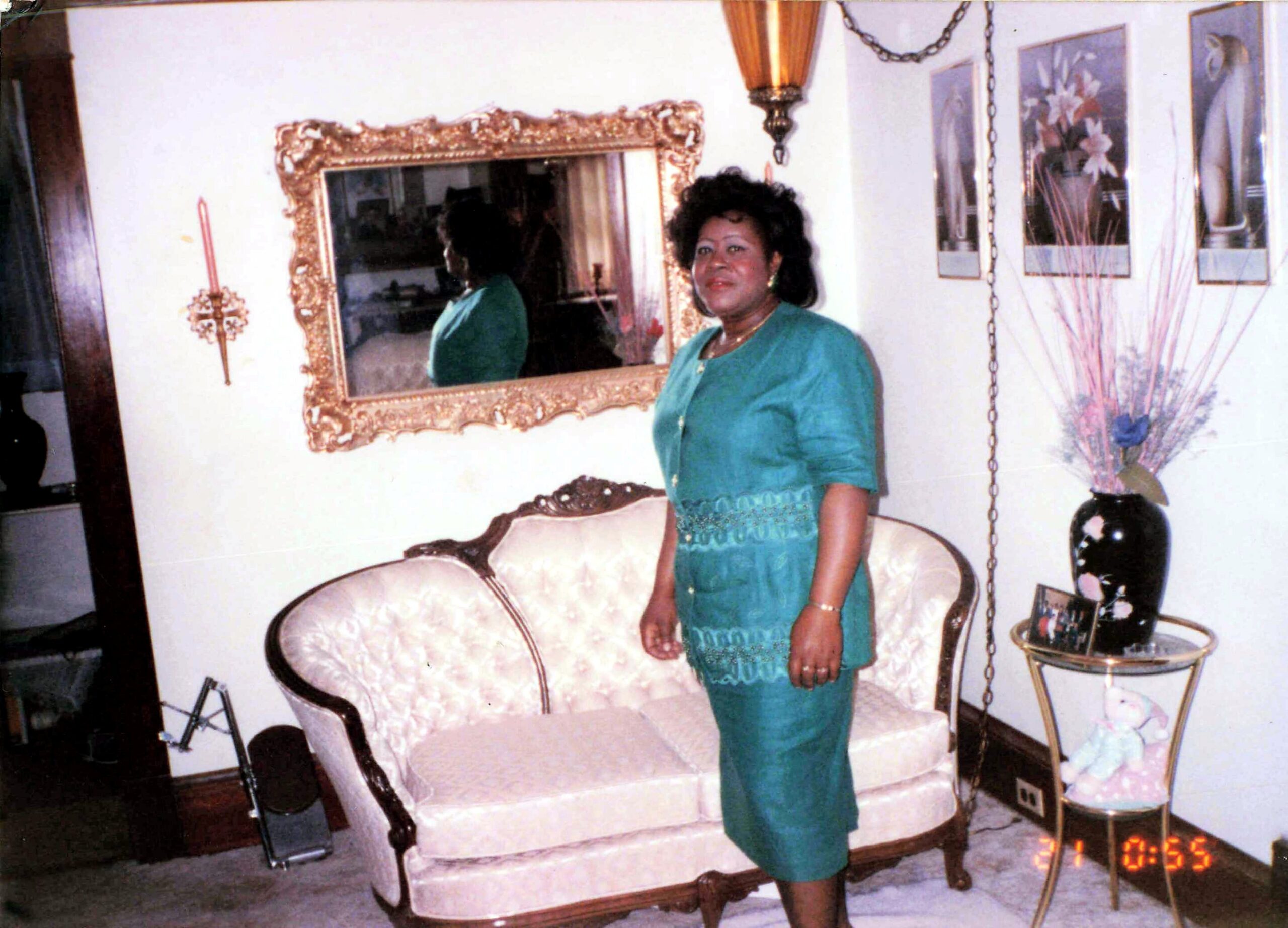 Woman in a teal dress standing beside a cream tufted sofa in a living room with a large ornate gold mirror.