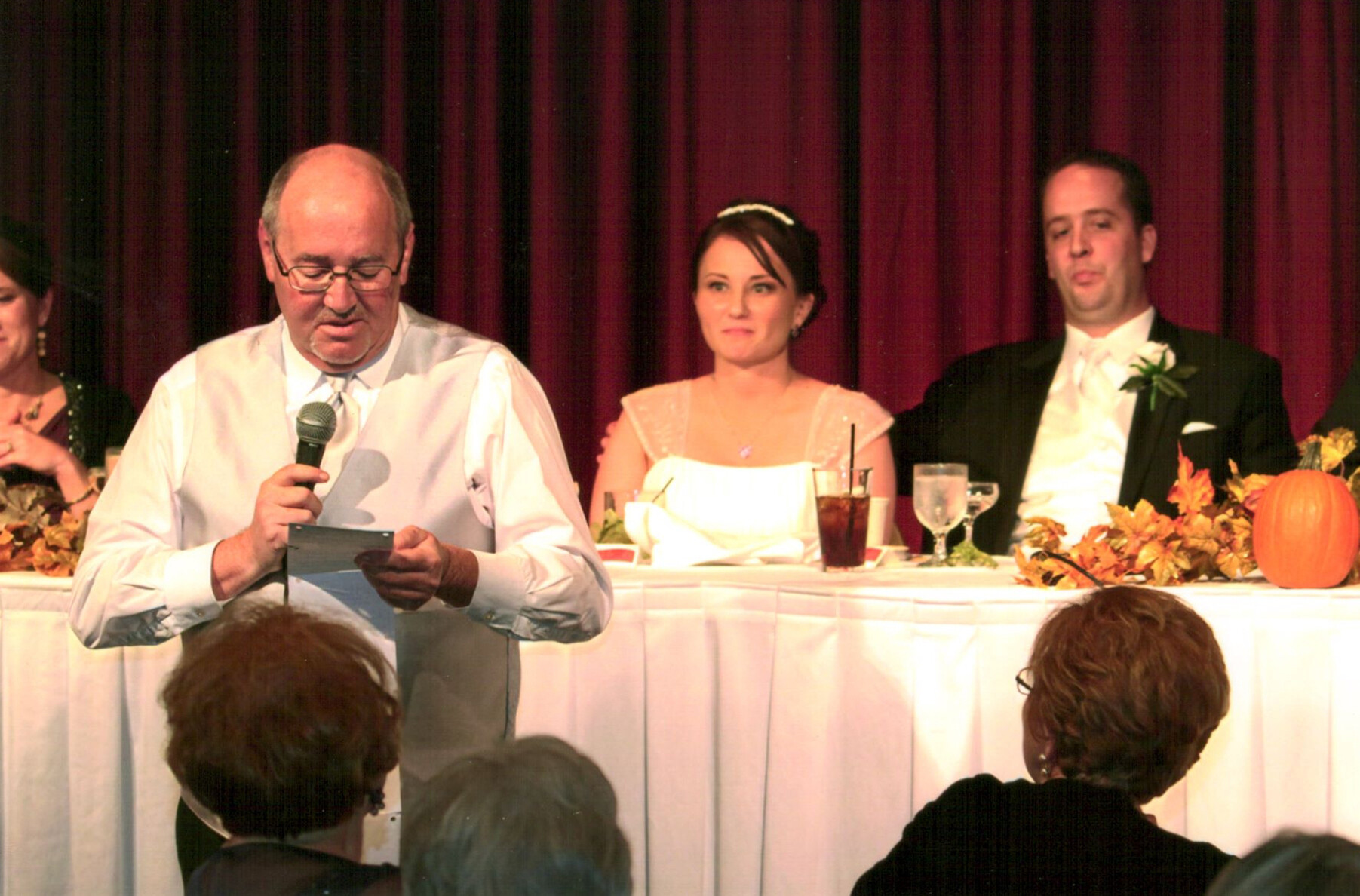 Man in a light vest speaks into a microphone at the head table during a wedding reception, with the bride and groom seated behind him against a burgundy curtain.