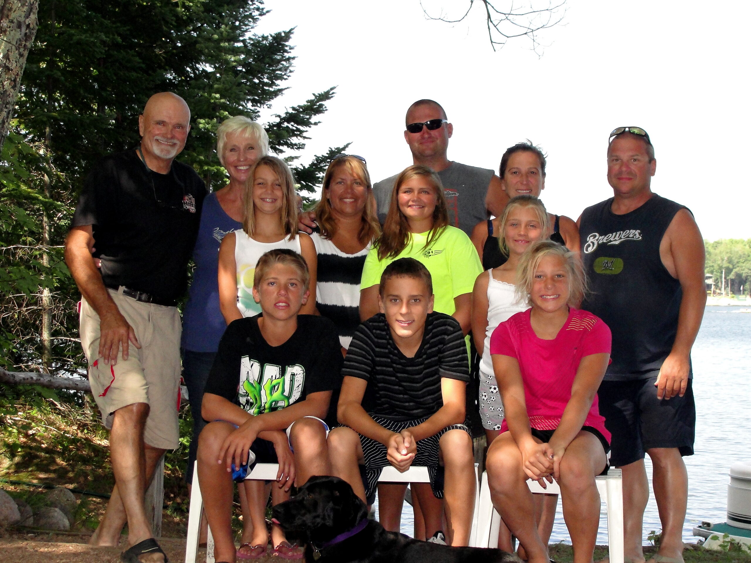 Group of adults and children posing together on a dock by a lake on a sunny day, smiling at the camera.