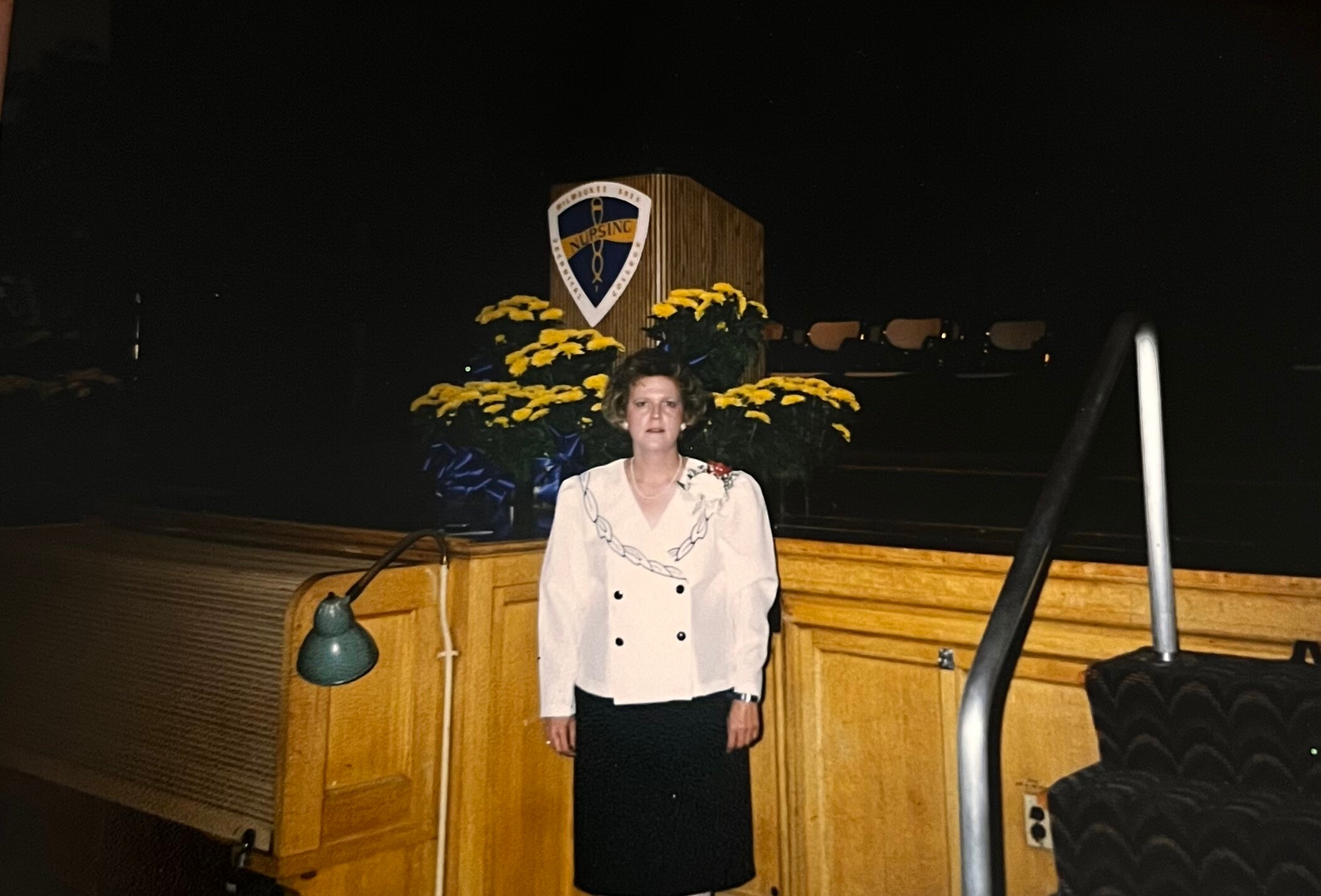 Woman standing at a wooden lectern adorned with yellow flowers and a shield-emblem behind her on stage.
