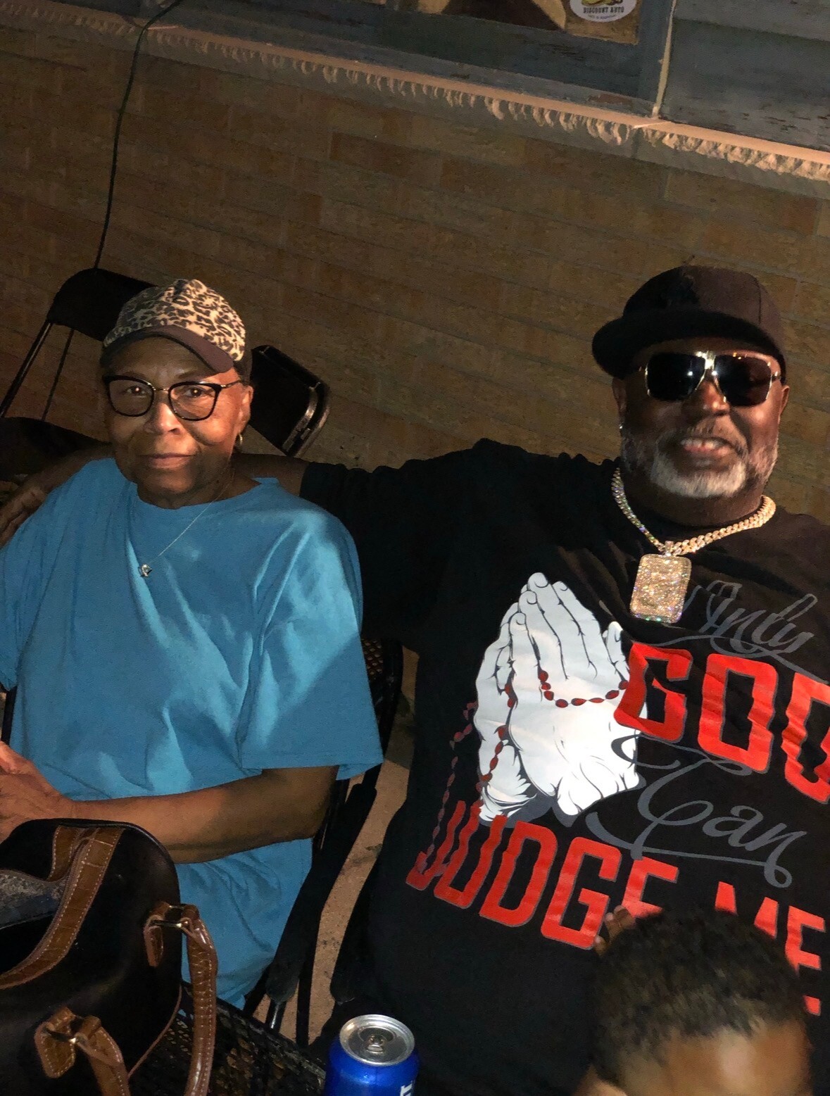 Two people posing together at night; the man on the right wears a cap, sunglasses, a large diamond pendant, and a black graphic shirt, while the woman on the left wears a blue shirt and a leopard-print cap at a brick background.