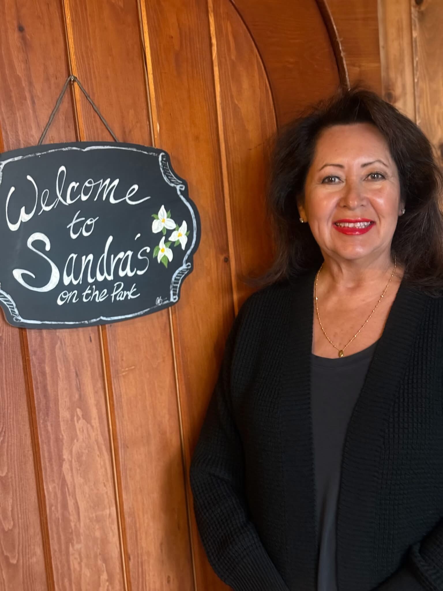Smiling woman in a black cardigan stands beside a wooden wall and a chalkboard sign reading, 'Welcome to Sandra's on the Park' with white flowers.