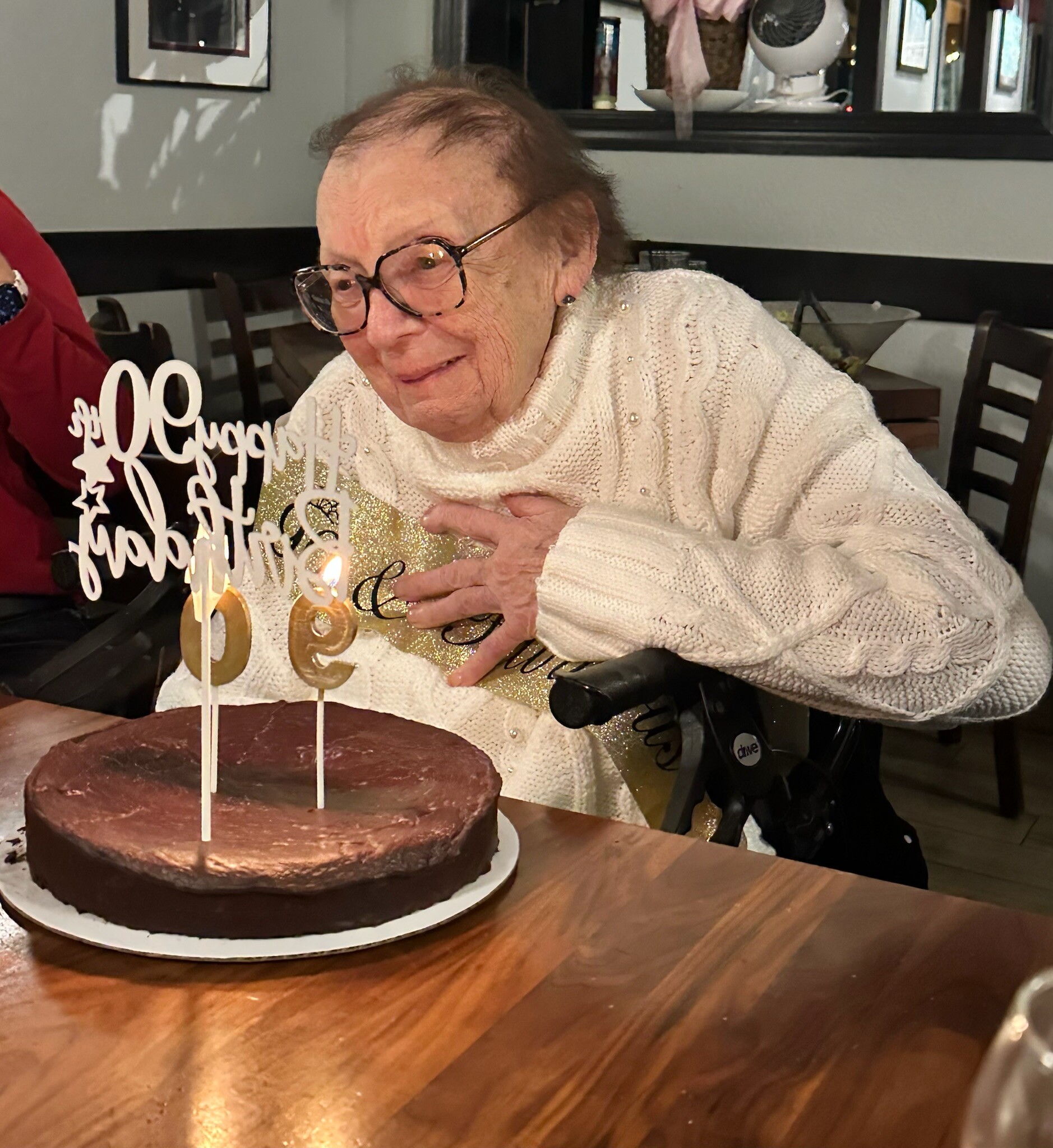 Smiling elderly woman in a white knit sweater sits in a wheelchair in front of a chocolate birthday cake with a gold 'Happy Birthday' topper and lit candles.