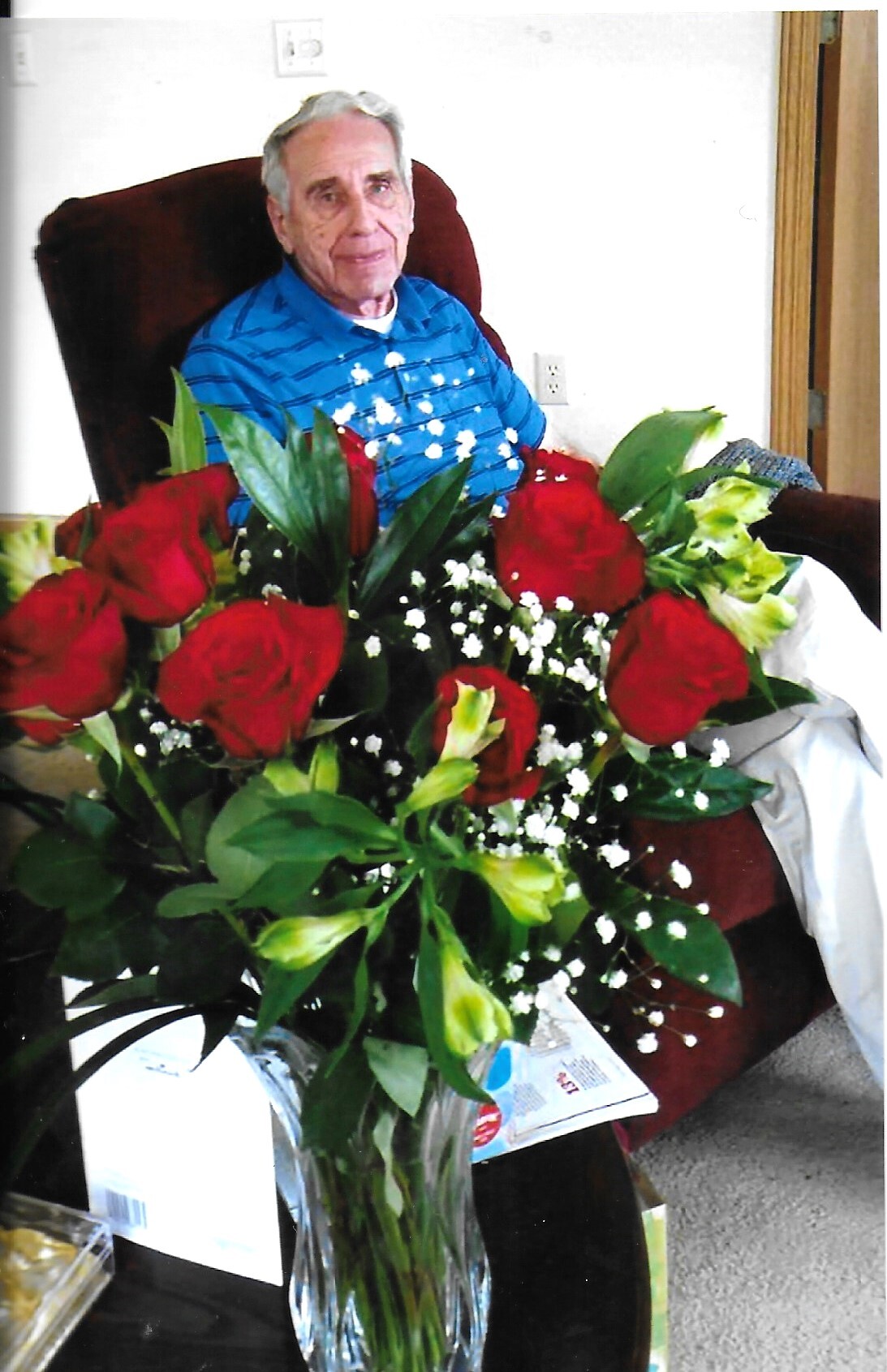 Elderly man in a blue striped shirt sits in a chair behind a tall vase of red roses and white baby's breath lilies.
