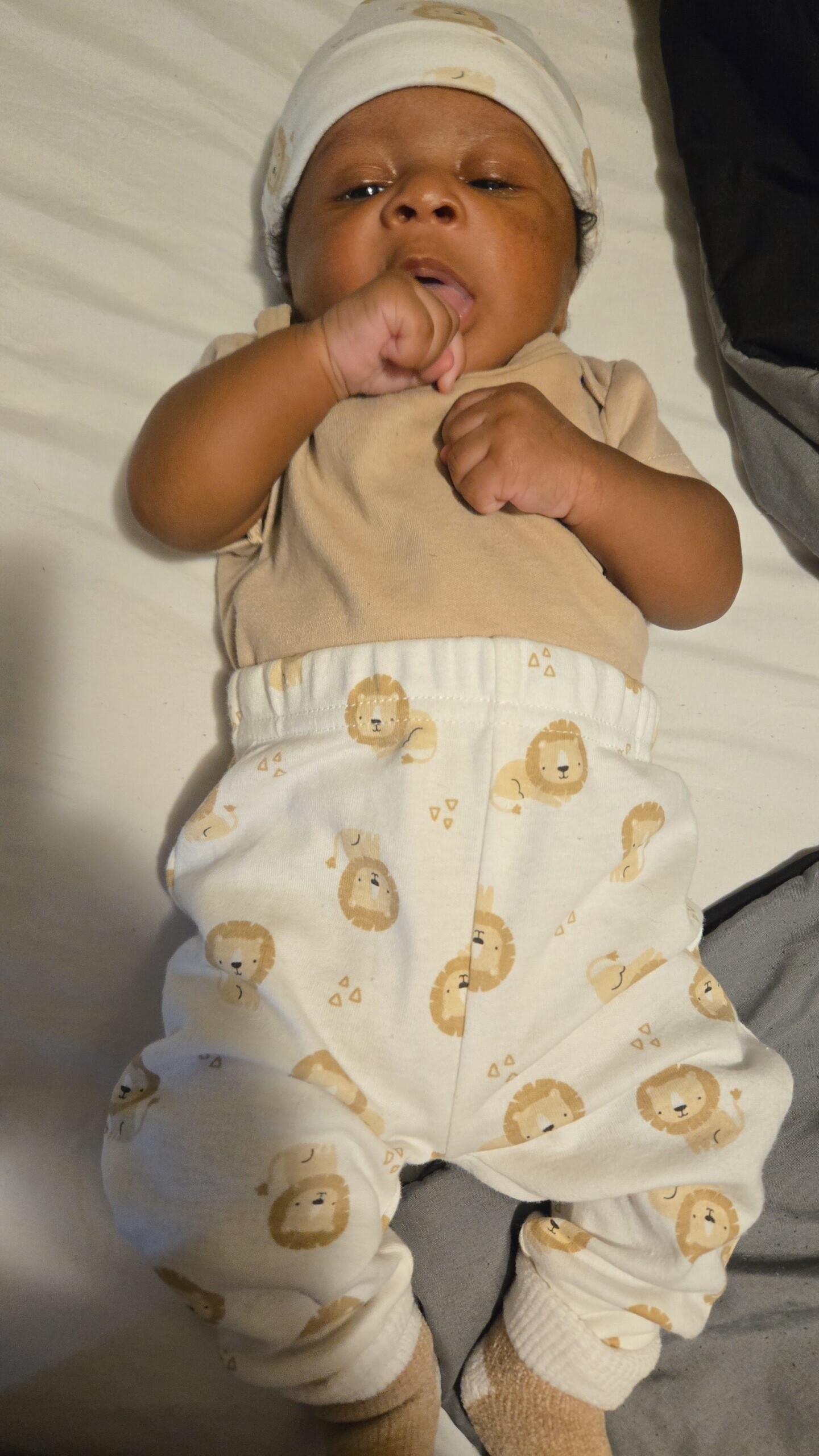 Baby lying on a bed in a beige shirt and white pants with lion prints, wearing a light hat.