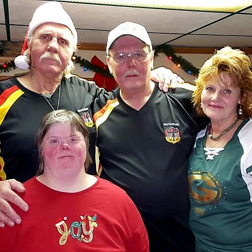 Group of four adults posing at a holiday gathering; two men in black shirts (one wearing a Santa hat, the other with a cap) stand behind a person in a red shirt, and a woman in a green top stands on the right.