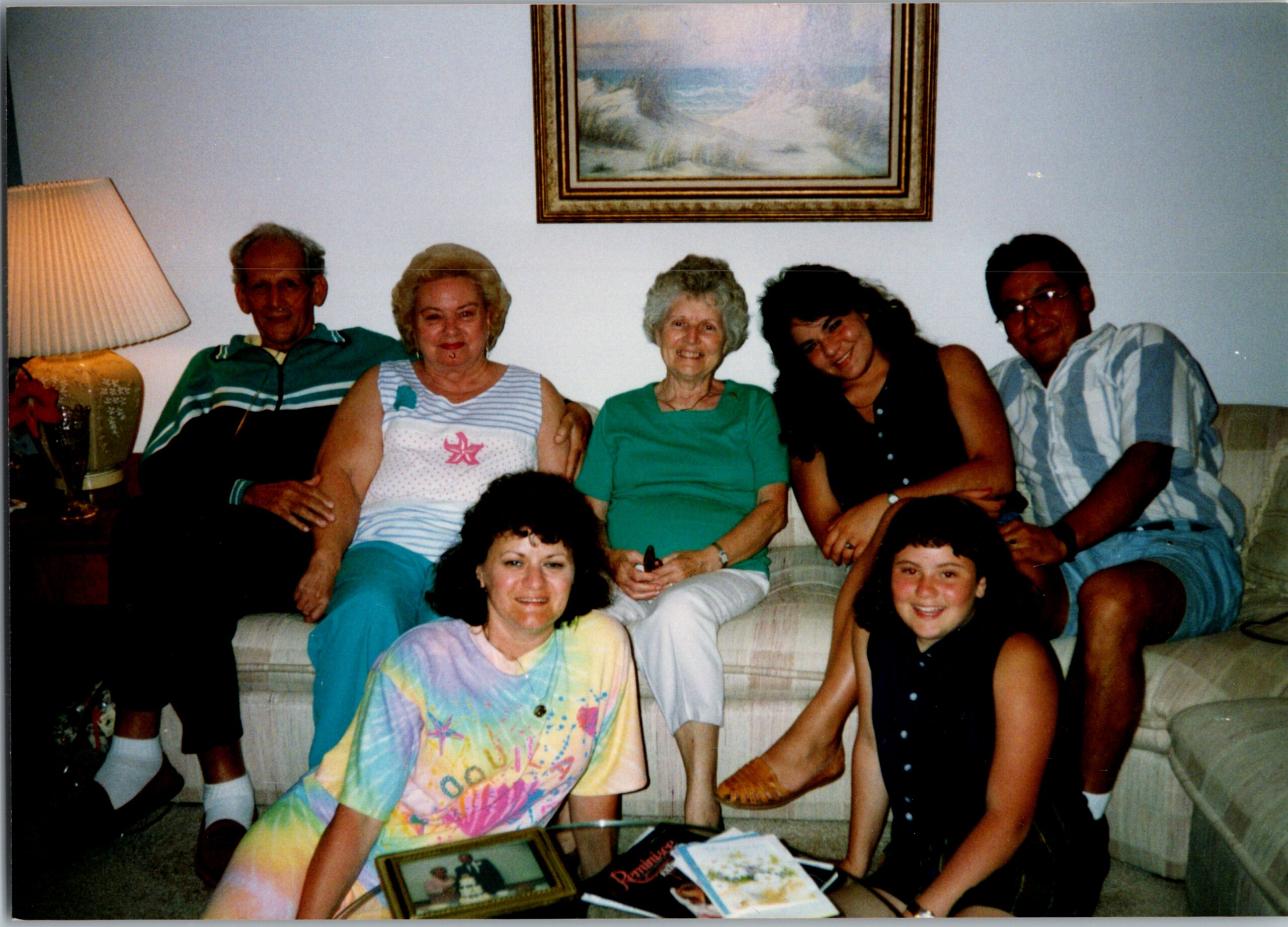 Group of six relatives posing on a living-room sofa, smiling for the camera; lamp on the left and a framed painting above.