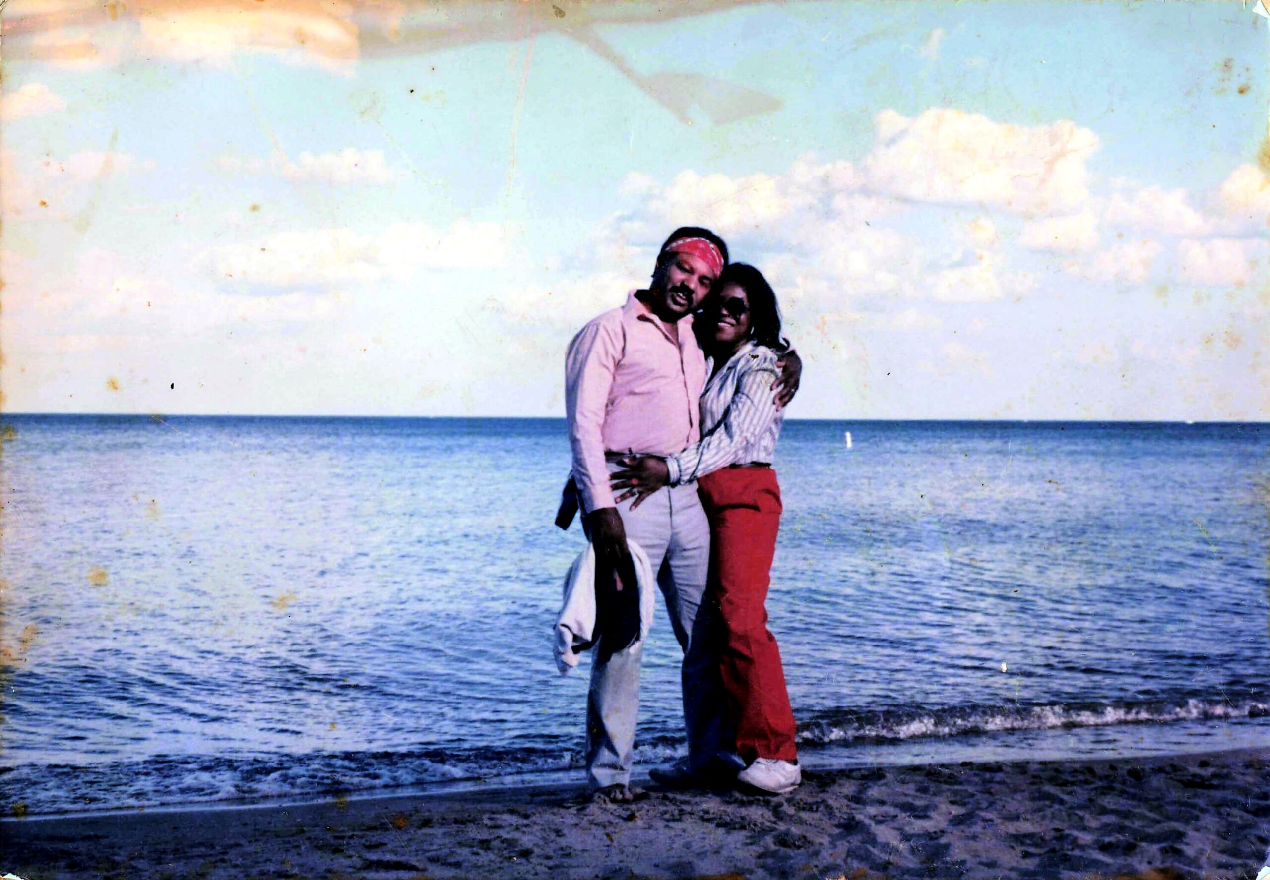 A couple hugs on a sandy beach with the calm sea and blue sky behind them.