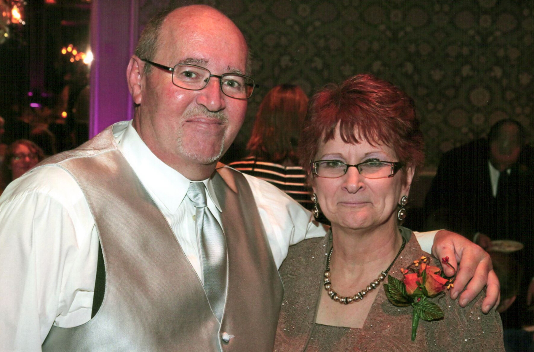 Smiling couple posing at a formal event: man in a beige vest and light tie, woman with short red hair wearing a sparkly dress and corsage, arms around each other