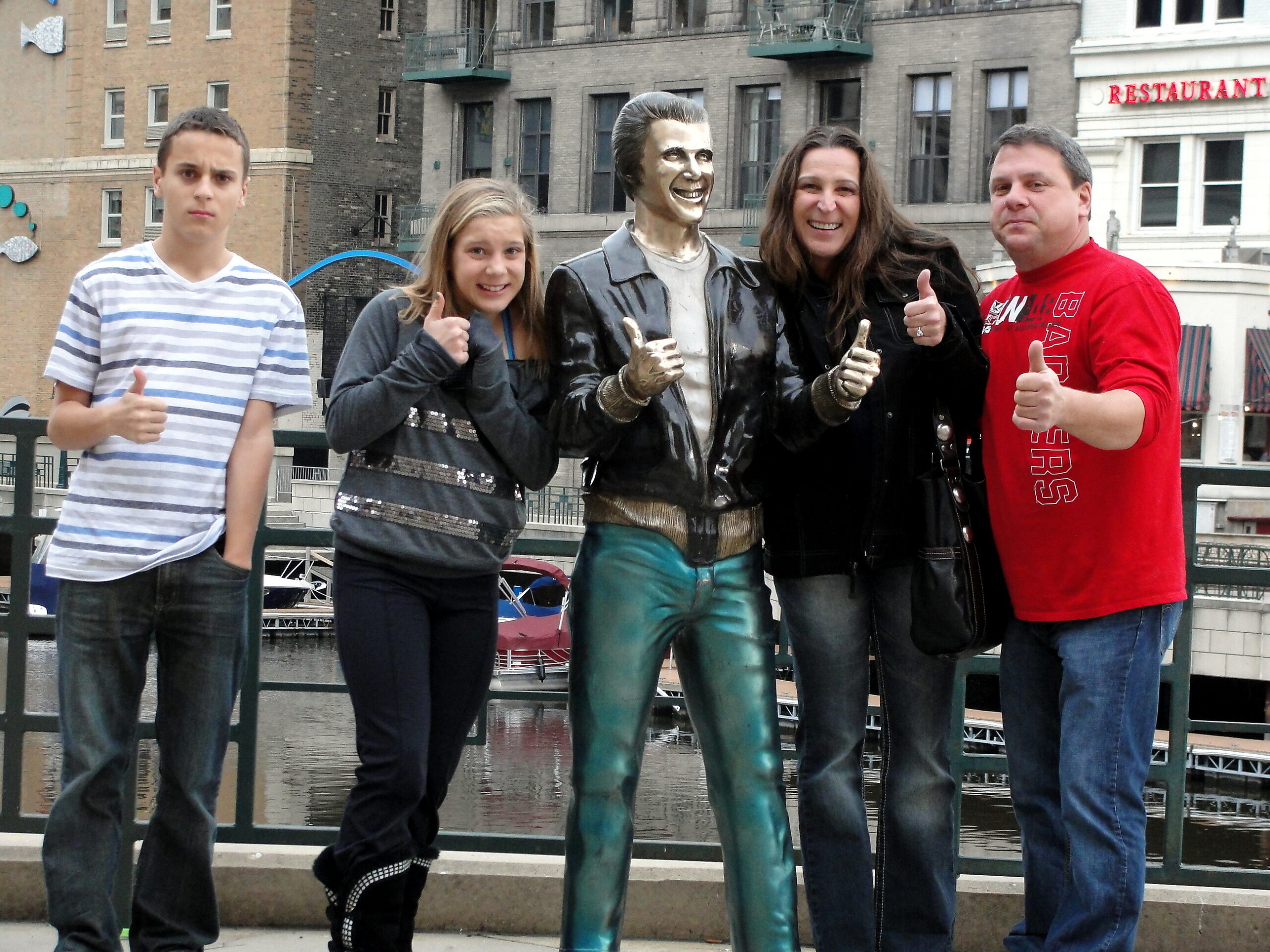 Five people posing on a riverside walkway with a shiny statue in the center, all giving thumbs up.