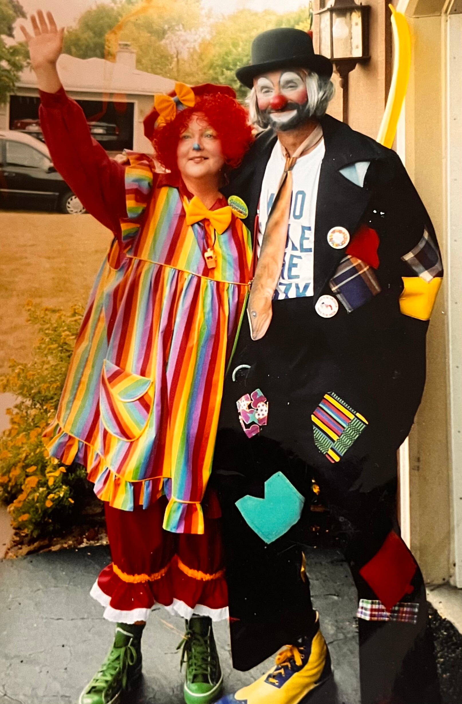 Two people in colorful clown costumes pose on a sidewalk; woman with a rainbow-striped dress and red wig waves beside a man in a black coat with white-painted clown makeup.