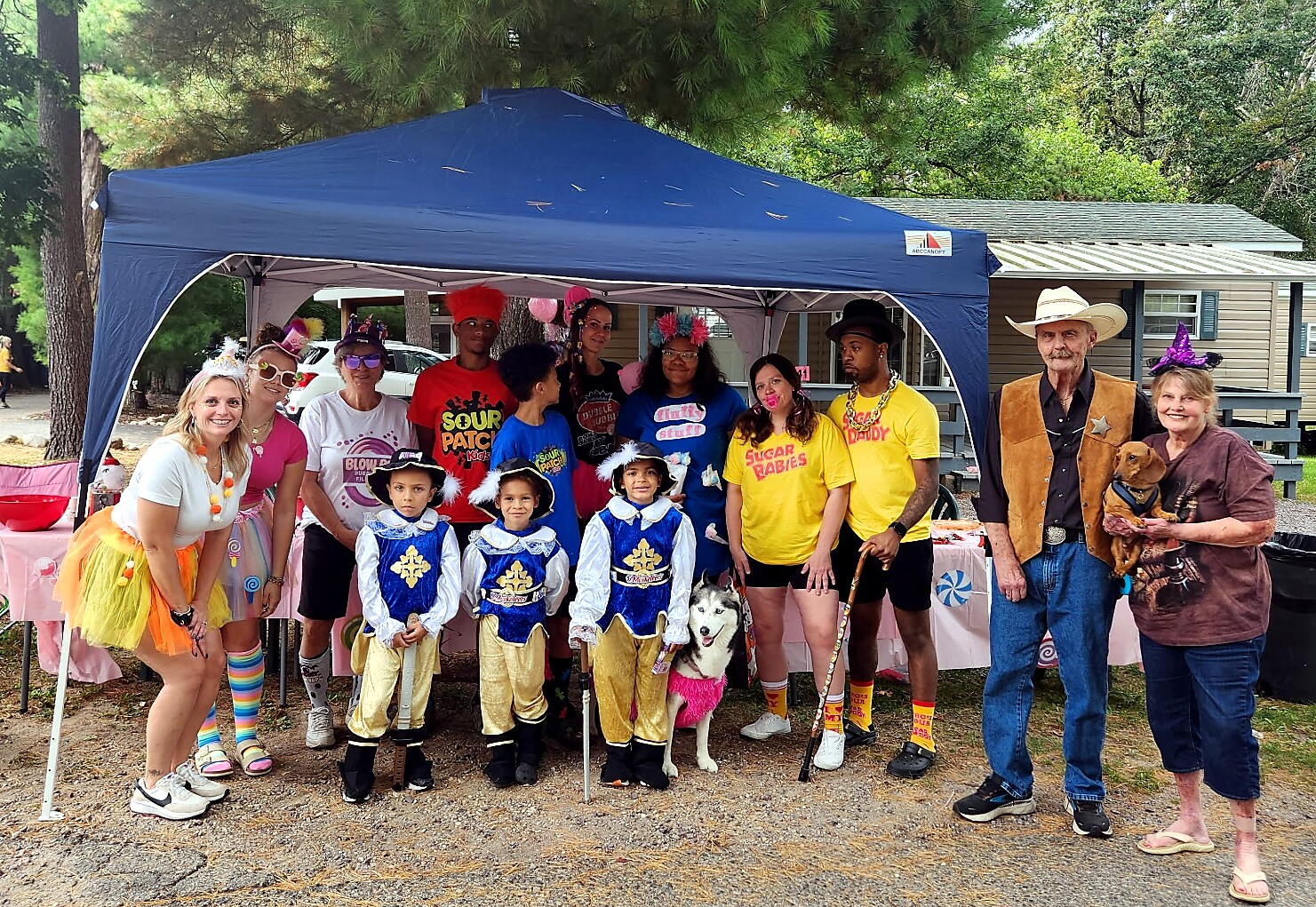 Group photo under a blue canopy at a costume party; children in blue prince outfits and adults in colorful costumes pose together outdoors under trees with a house in the background