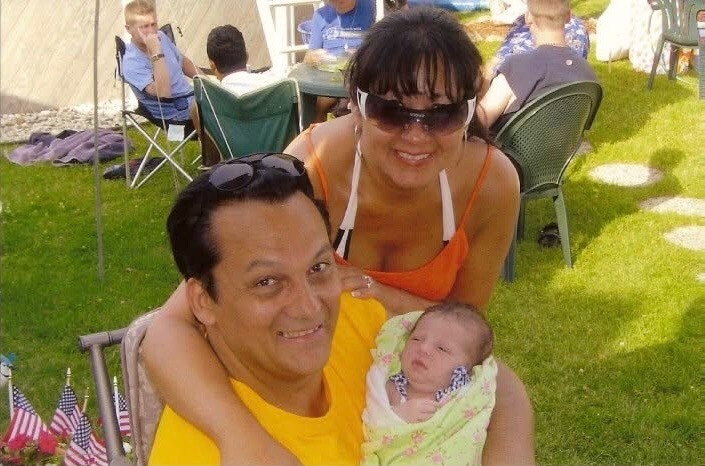 Man smiles while holding a newborn wrapped in a light blanket at an outdoor family gathering; a woman in sunglasses leans over them from behind.