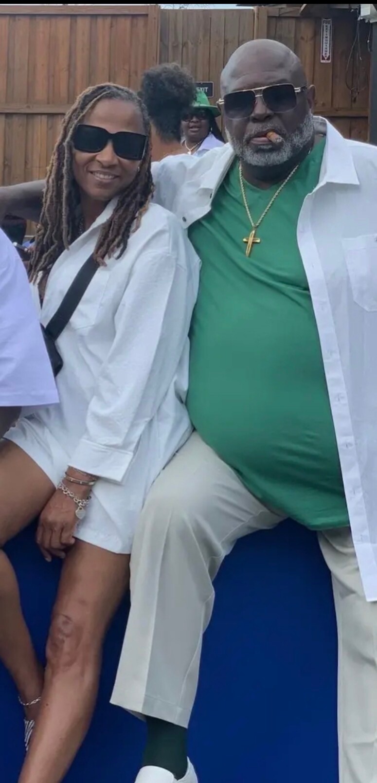 Two adults in white coats posing outdoors; woman with sunglasses and dreadlocks, man with green shirt, gold cross, and a cigar nearby.