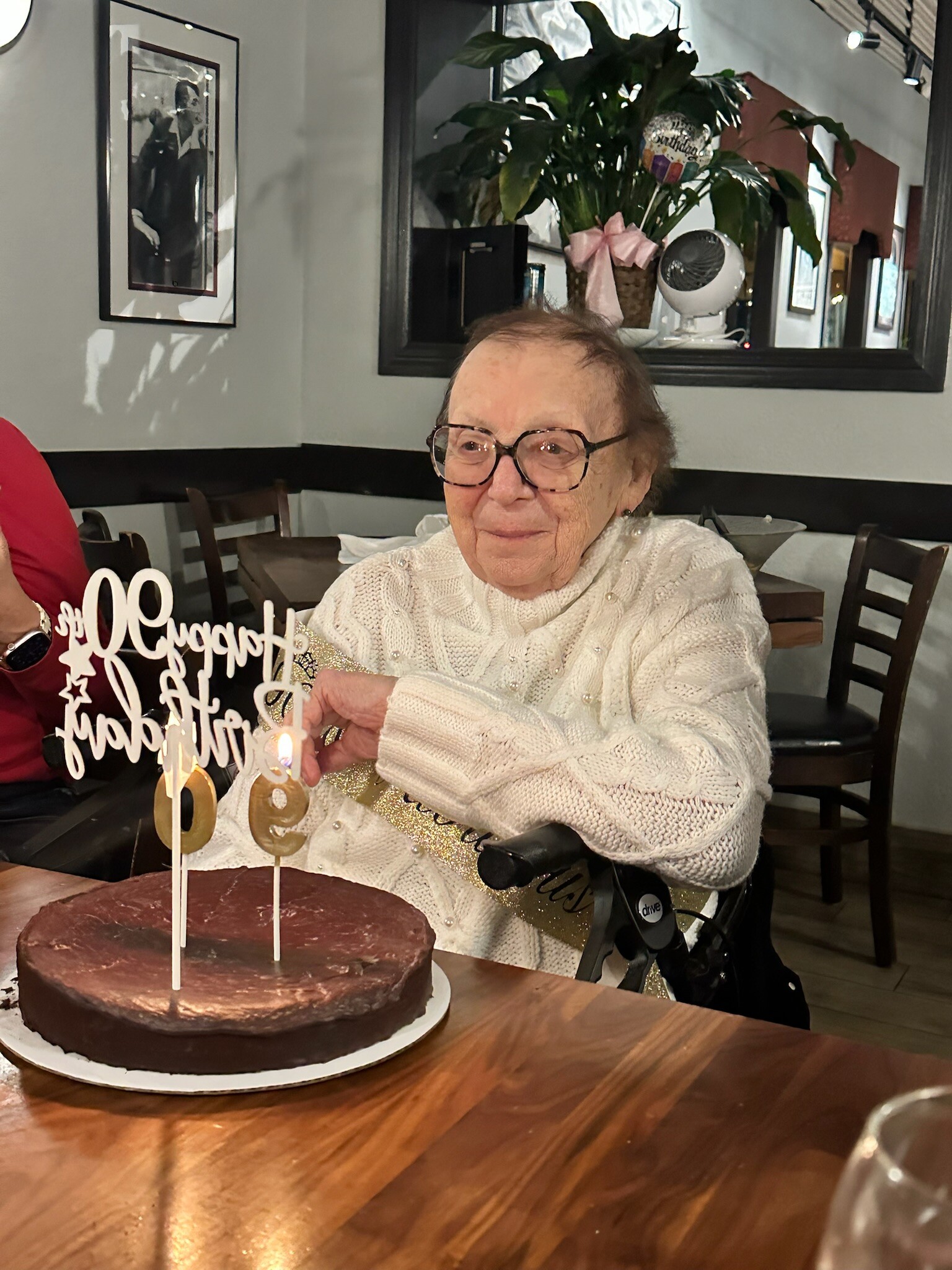 Elderly woman in a white sweater smiling at a chocolate birthday cake with candles; seated in a wheelchair at a restaurant table.