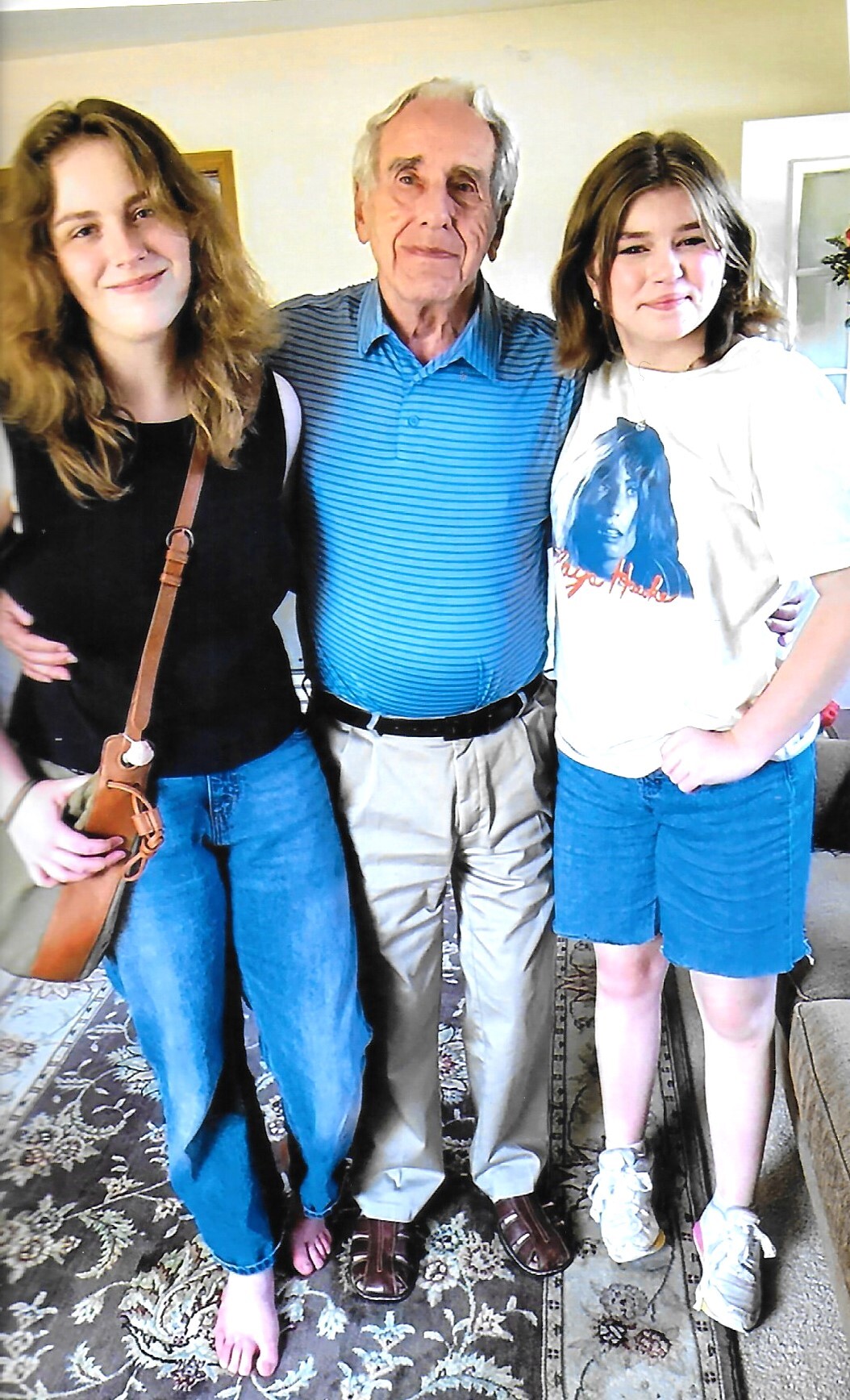 Three people stand together indoors, smiling: an older man in a blue striped polo flanked by two young women in casual outfits.