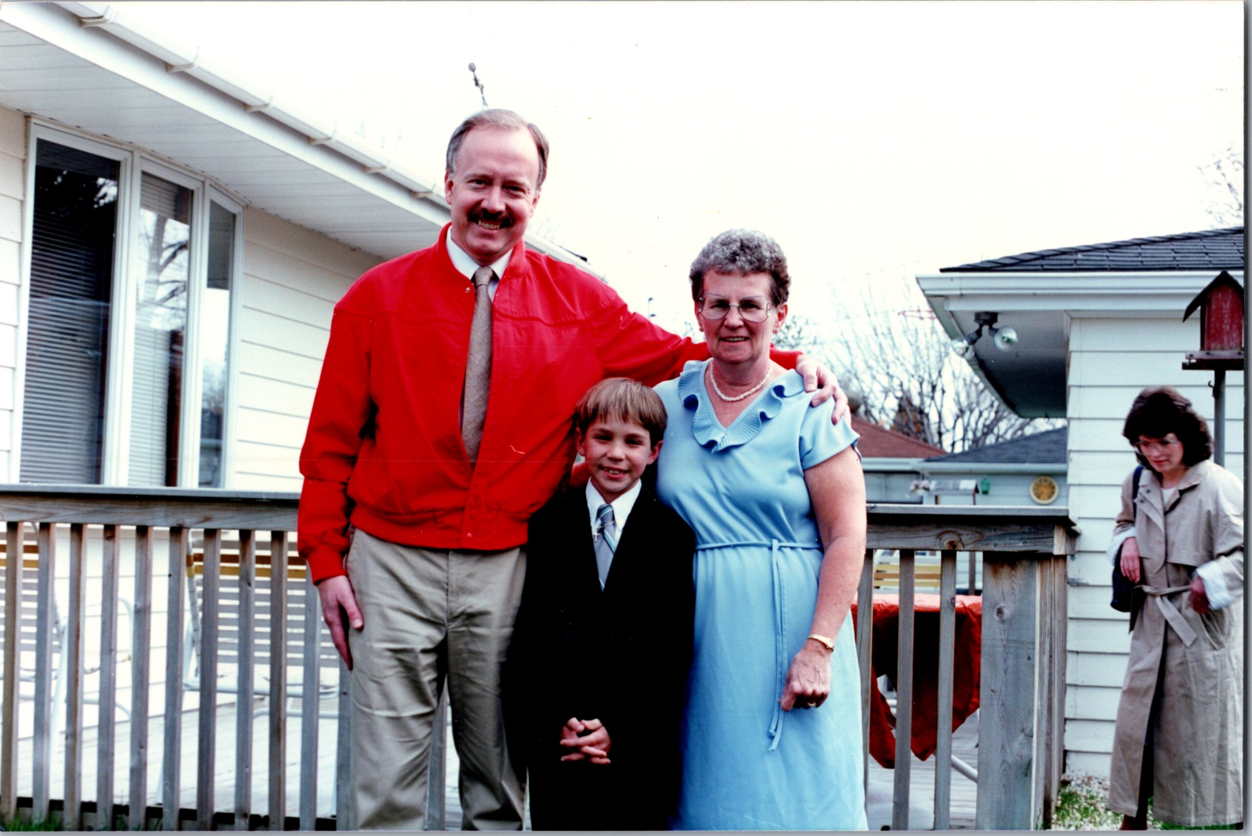 Smiling man in a red jacket and woman in a blue dress pose with a boy in a suit on a wooden deck outside a house.