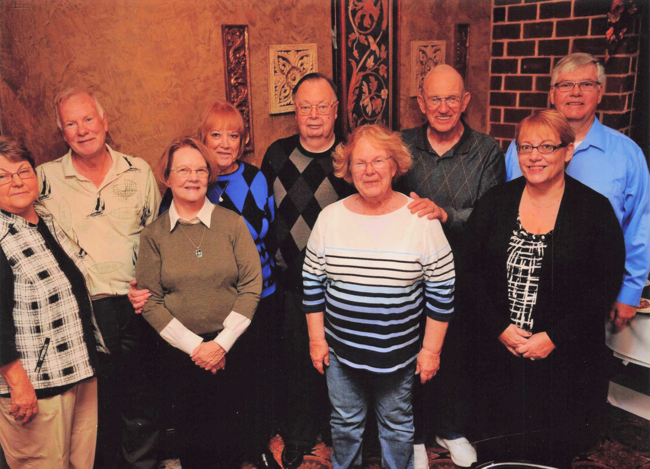 Group portrait of eight adults standing together indoors, smiling, in casual clothing against a textured wall and decorative panels.