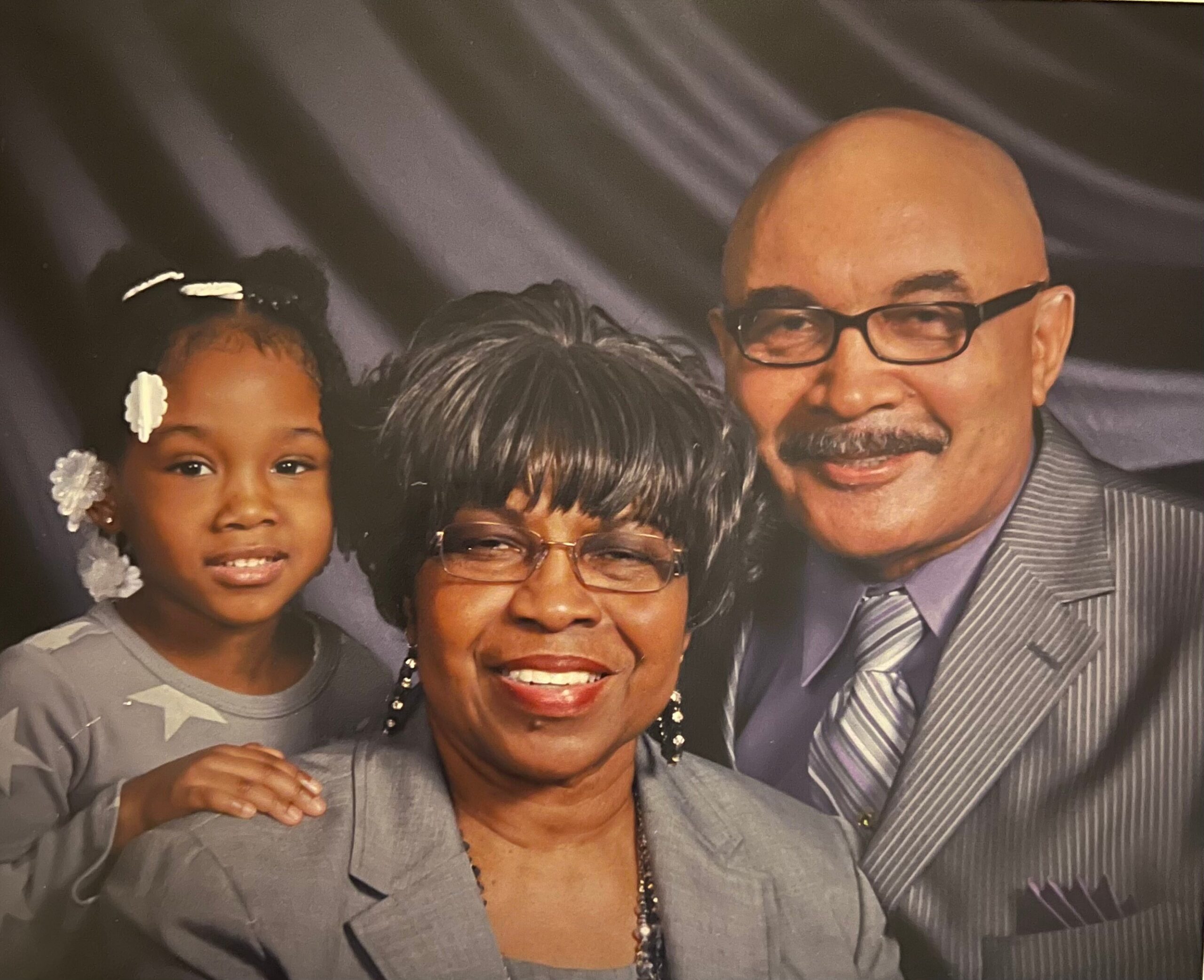 Three-generation family portrait: grandmother center, man wearing glasses to the right, and a young girl on the left smiling.
