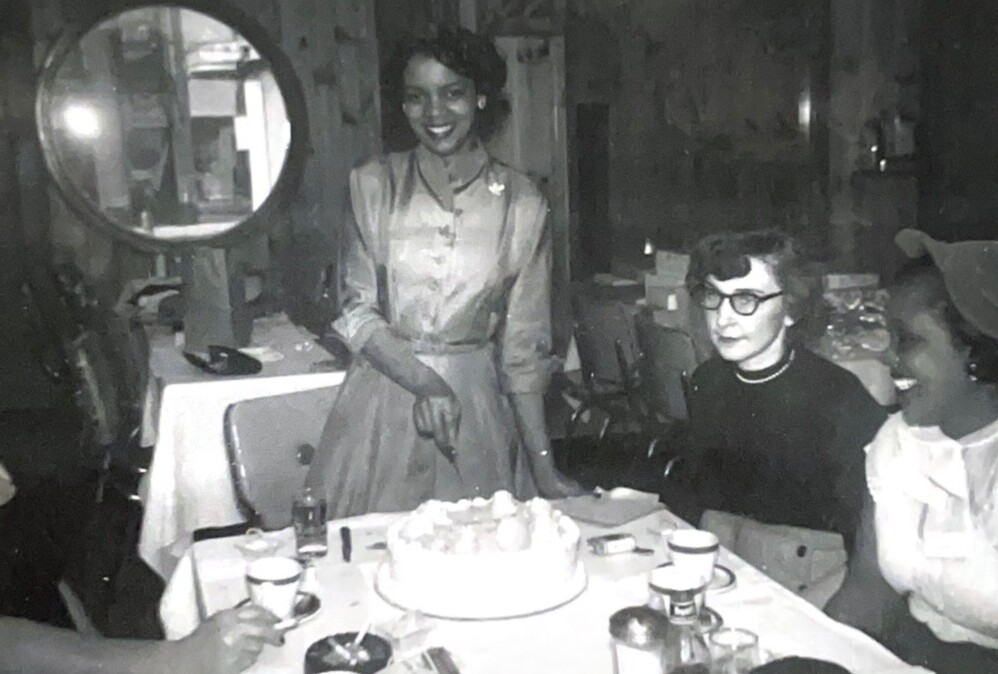 Black-and-white photo of a woman in a vintage dress standing beside a round cake on a table at a birthday party, with guests seated nearby.