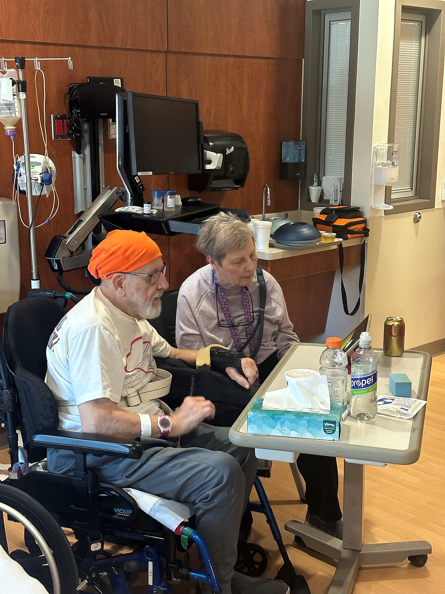 Two elderly people in a hospital room, one in a wheelchair with an orange hat, the other seated beside at a table with drinks and a tissue box nearby.