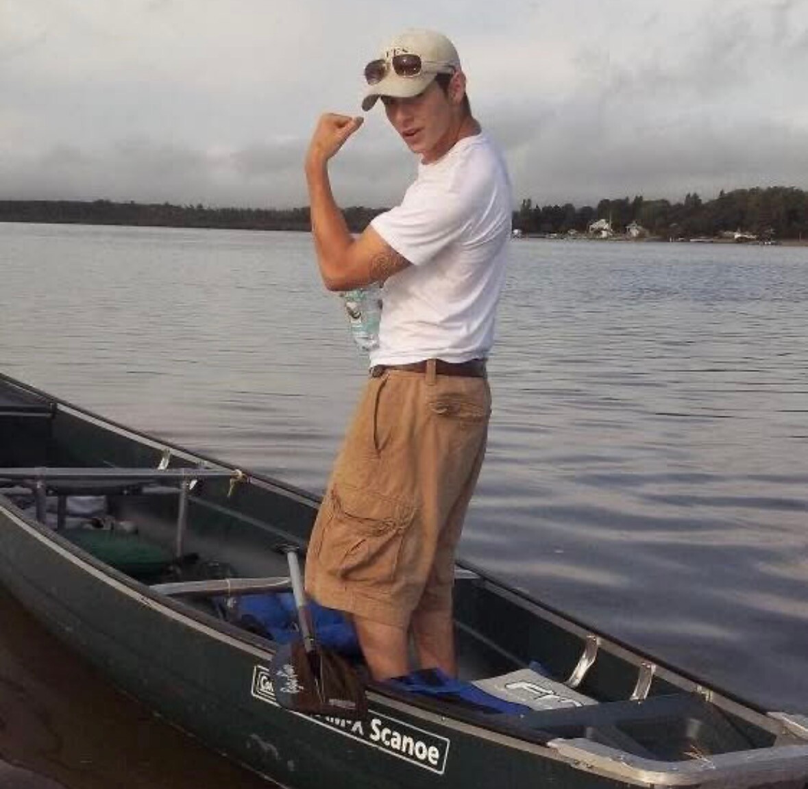 Man standing in a canoe on a calm lake, flexing his arm toward the camera, wearing a white t-shirt, tan cargo shorts, and a beige cap with sunglasses.