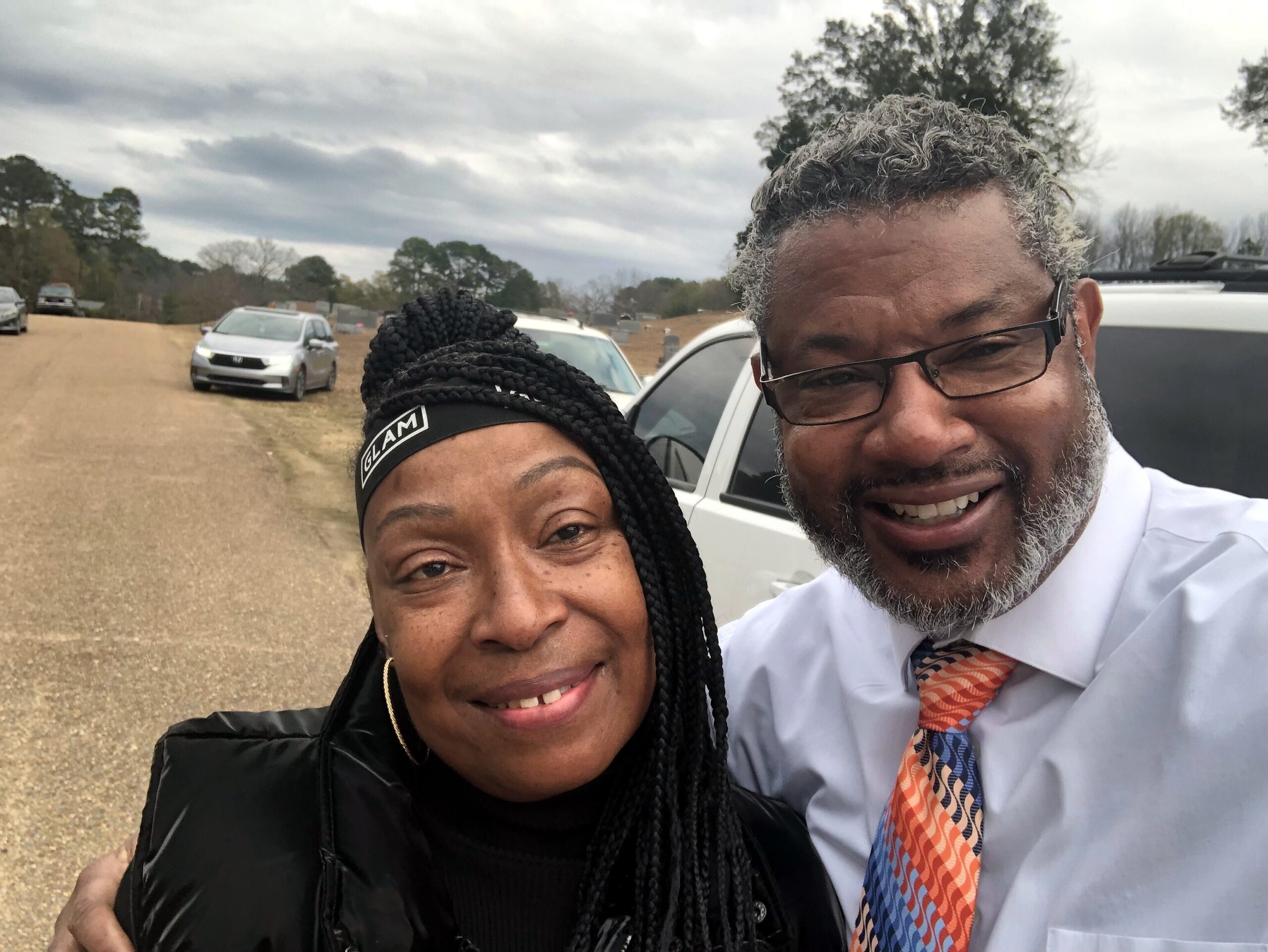 Two people smile for a selfie outdoors on a dirt road with parked cars in the background and a cloudy sky above.