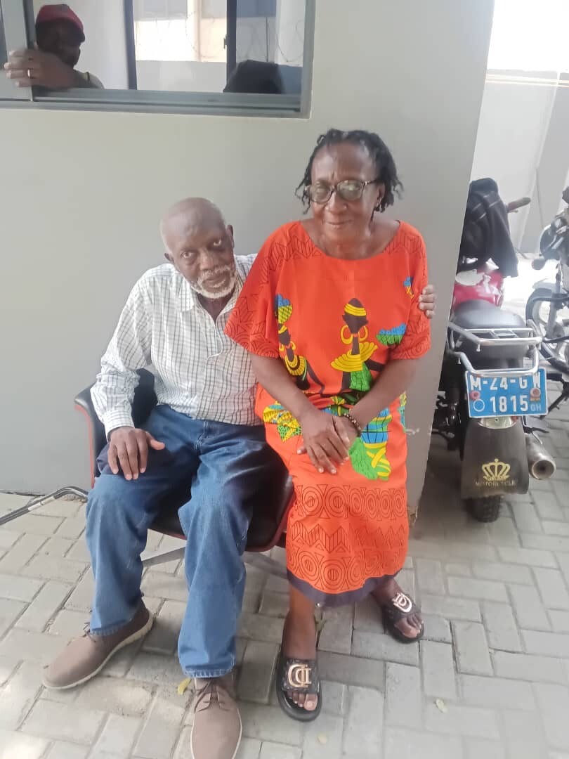 Elderly couple sitting together outside: man with white beard in a checkered shirt and jeans, woman in a bright orange patterned dress smiling beside him. A scooter with a blue license plate is in the background.