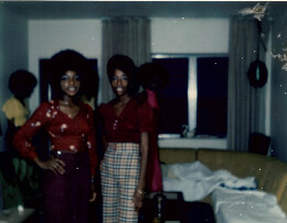 Two women pose in a living room wearing 1970s outfits; red tops, one in a purple skirt, the other in plaid pants, with a sofa behind.