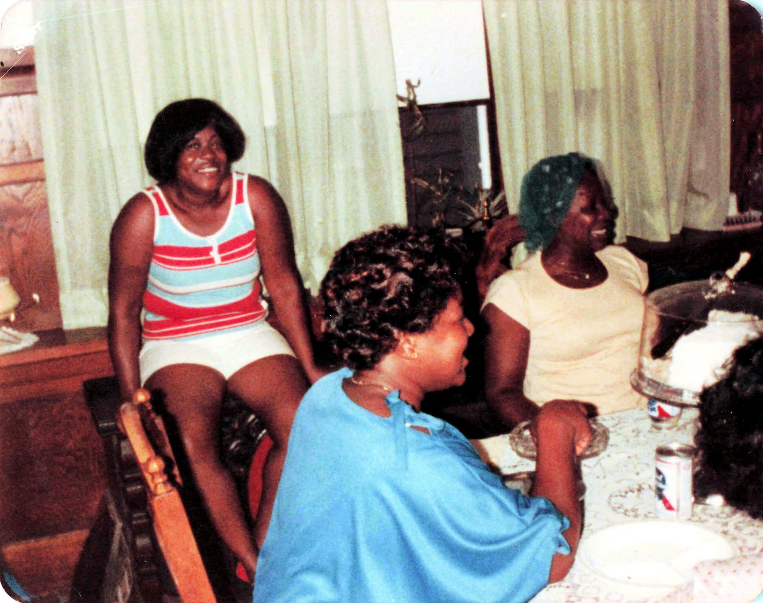 On a sunny indoor gathering, a smiling woman in a colorful striped swimsuit sits among friends and family beside a window with pale curtains.