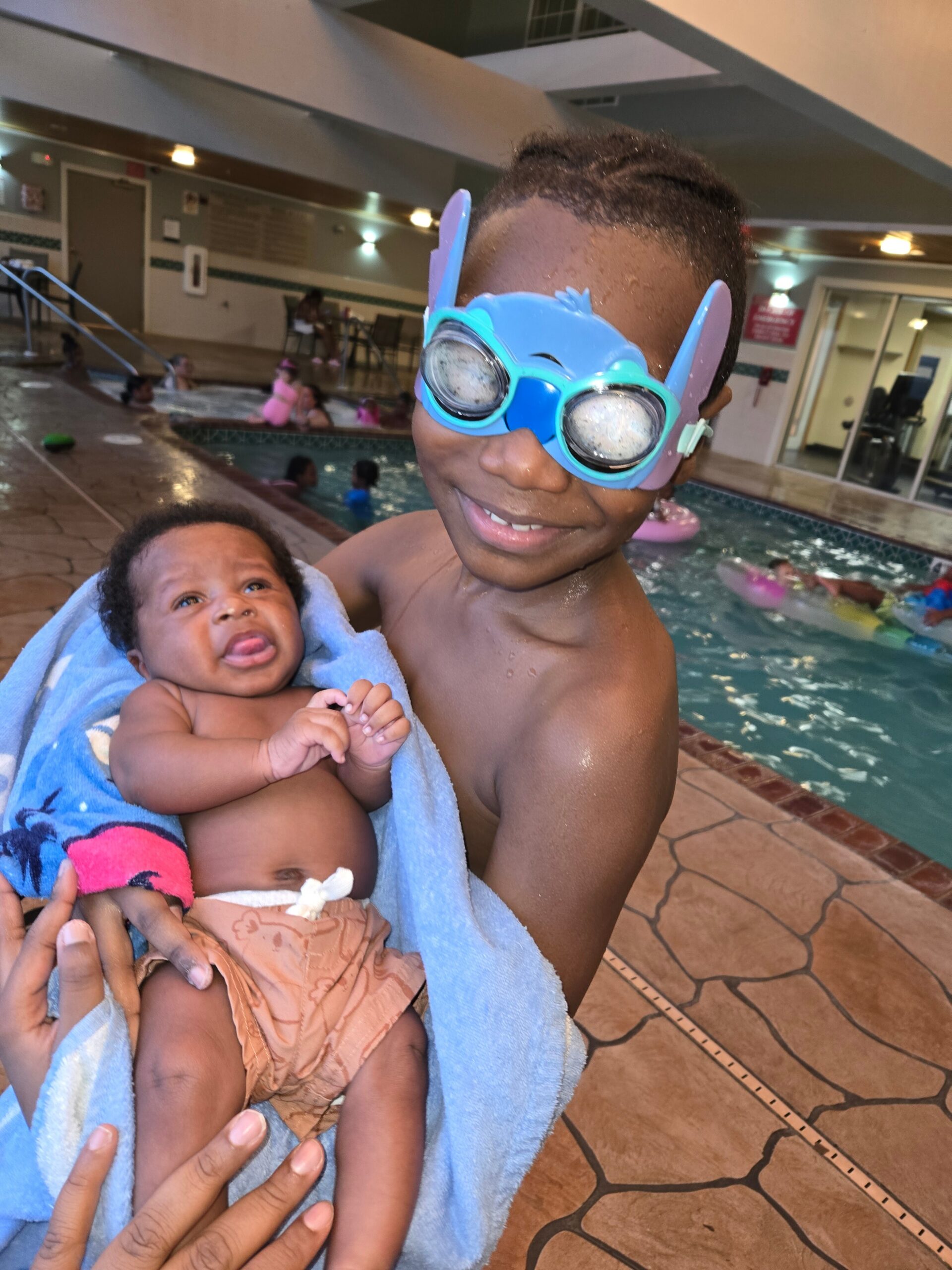 Boy wearing blue swim goggles smiles while holding a baby wrapped in a blue towel by an indoor pool, with other swimmers in the background.