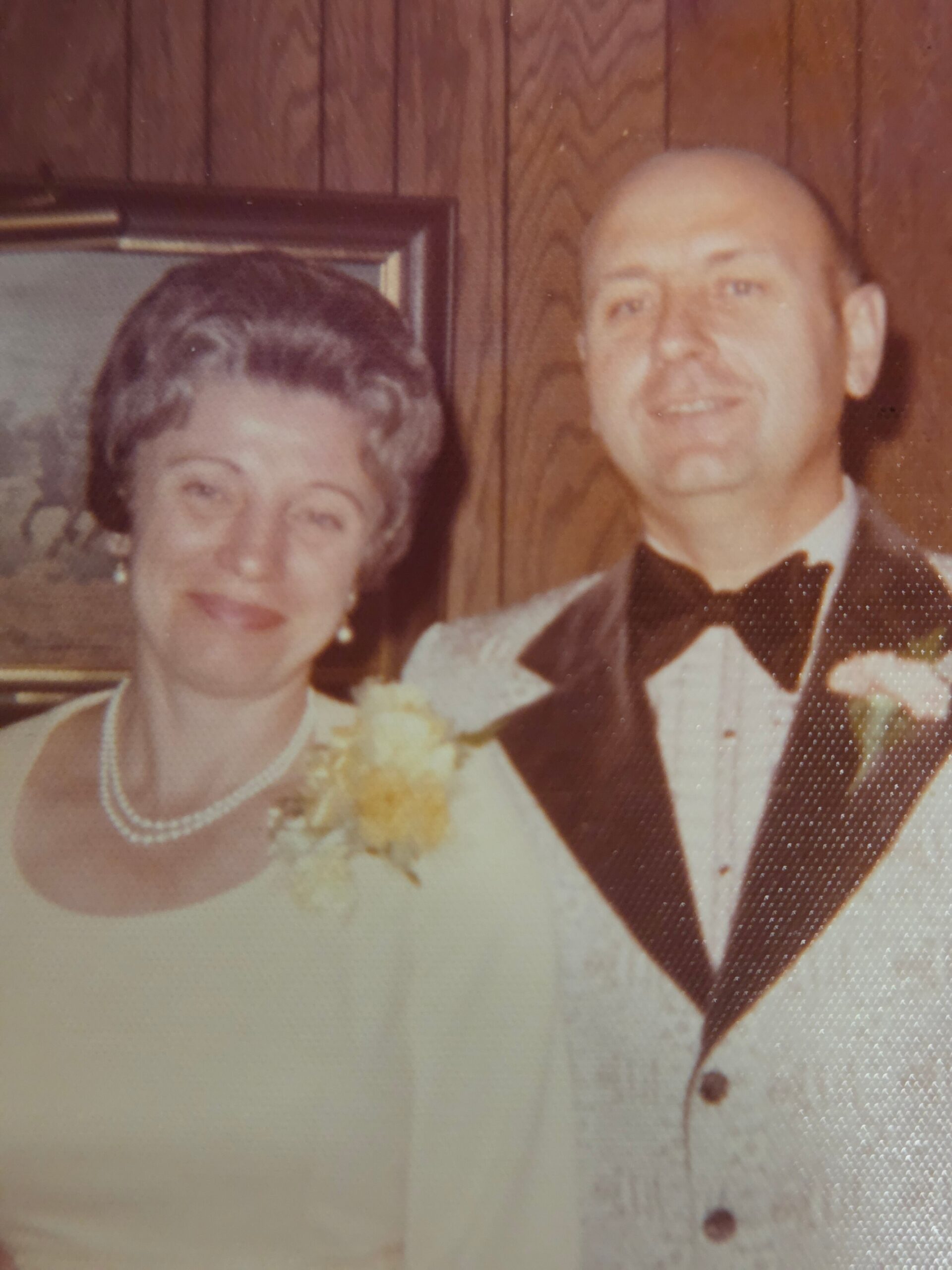 An elderly couple dressed in formal attire posing for a wedding photo; the woman wears a pearl necklace and a corsage, the man in a tuxedo with a bow tie, standing in front of wood-paneled walls.