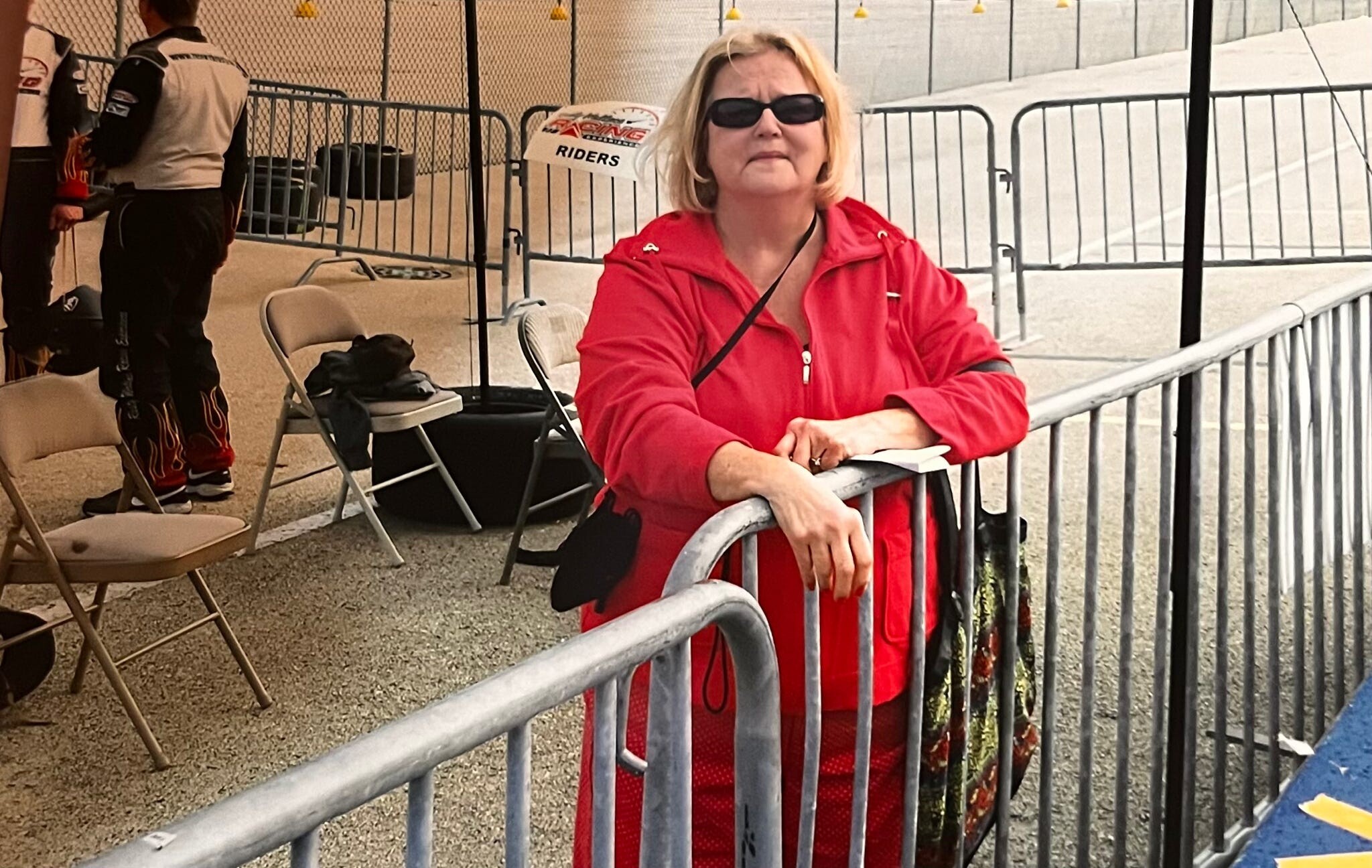 Woman with blonde hair and sunglasses wearing a bright red jacket leans on a metal barrier at an outdoor event, with empty chairs and a fenced area in the background.